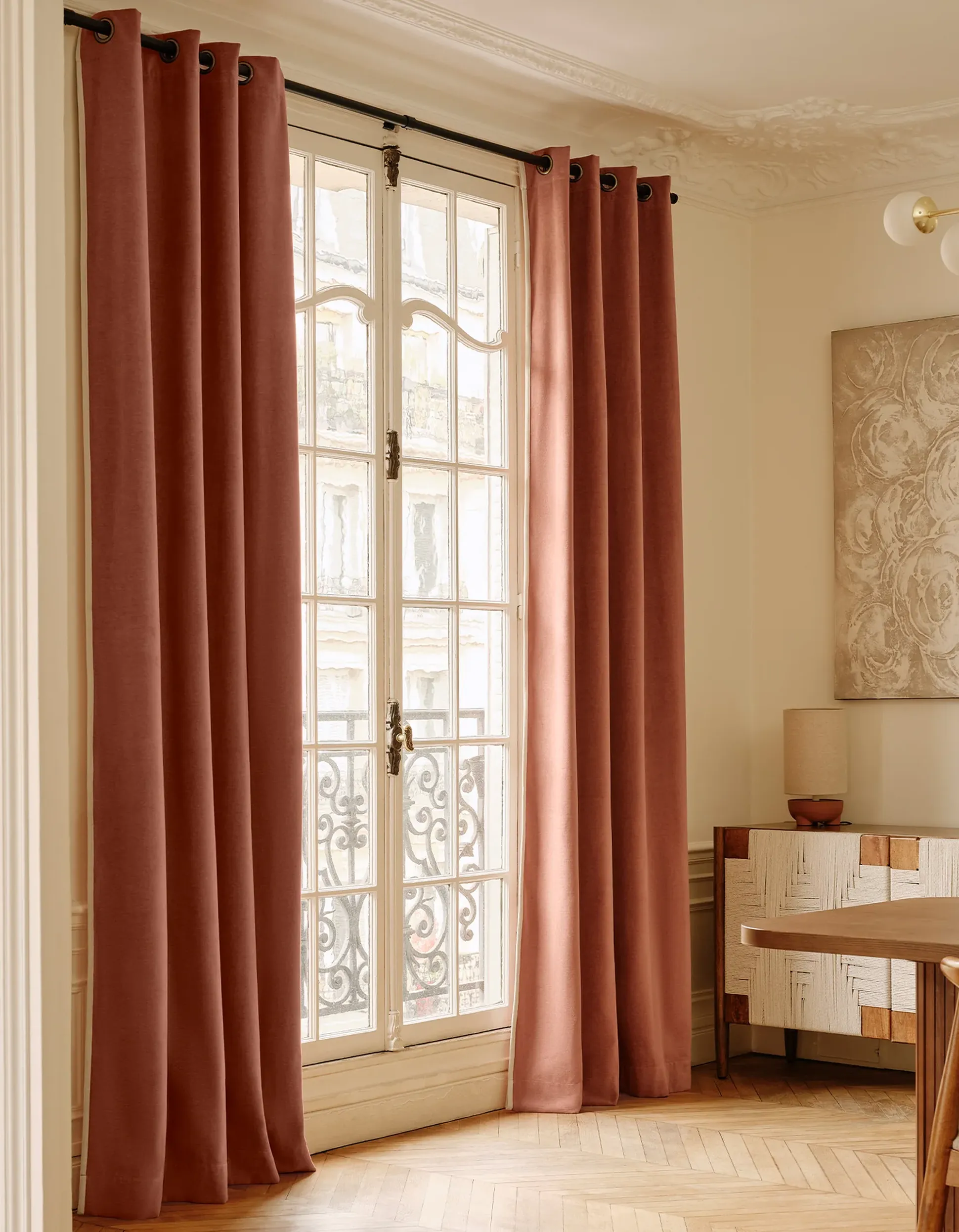 Chambre élégante avec de grands rideaux roses sur une fenêtre à cadre blanc, des meubles en bois à motifs et un sol en parquet. Décoration murale et éclairage discrets.