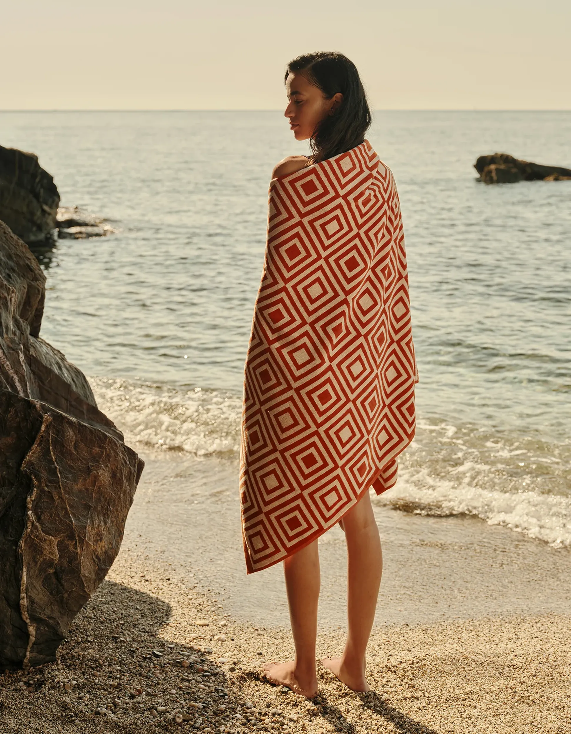Une personne enveloppée dans une serviette à motifs se tient debout sur une plage rocheuse, contemplant la mer calme sous un ciel dégagé.