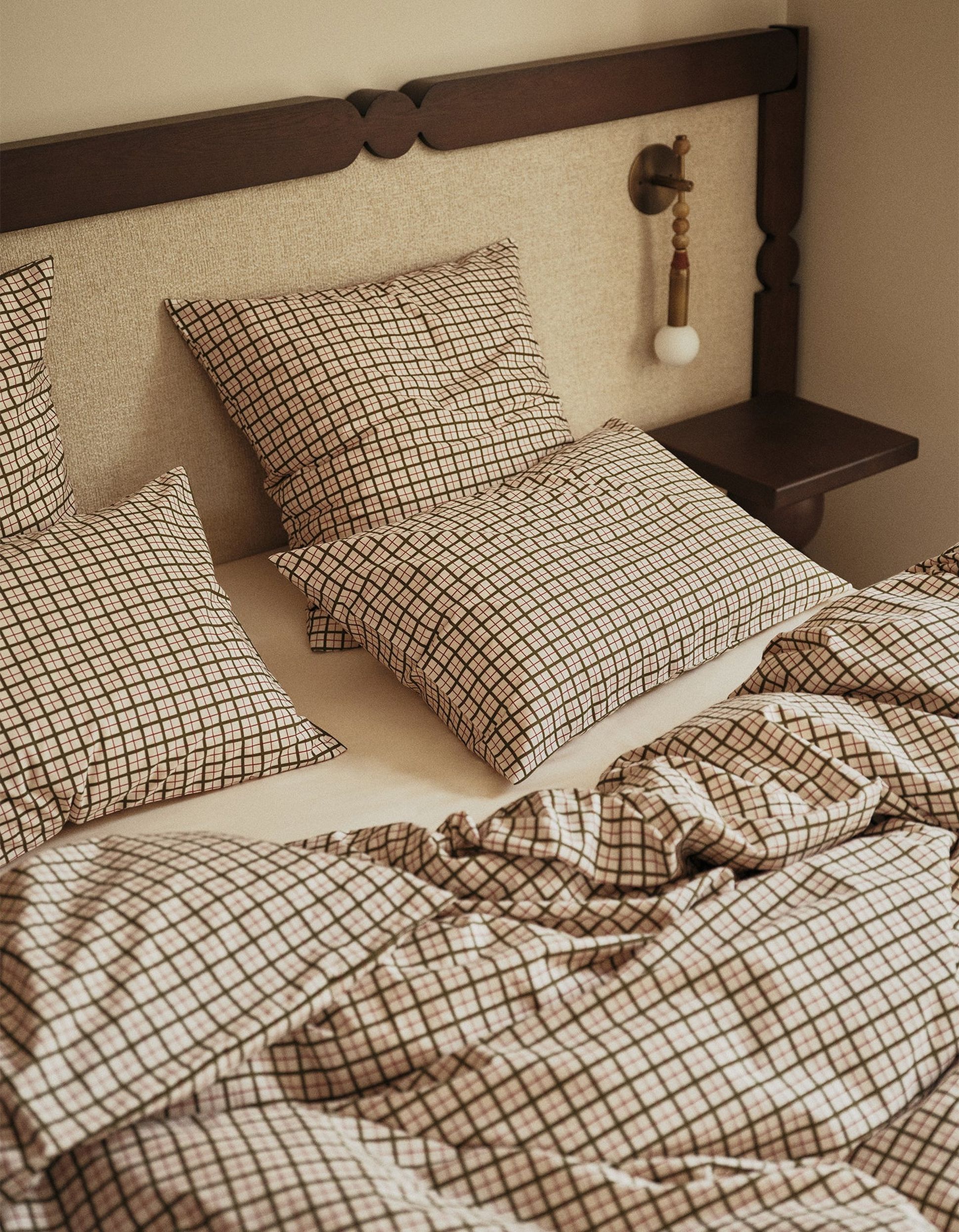 Un lit douillet avec des oreillers et une couette à carreaux, une table de chevet en bois et une tasse en céramique posée sur un drap blanc.