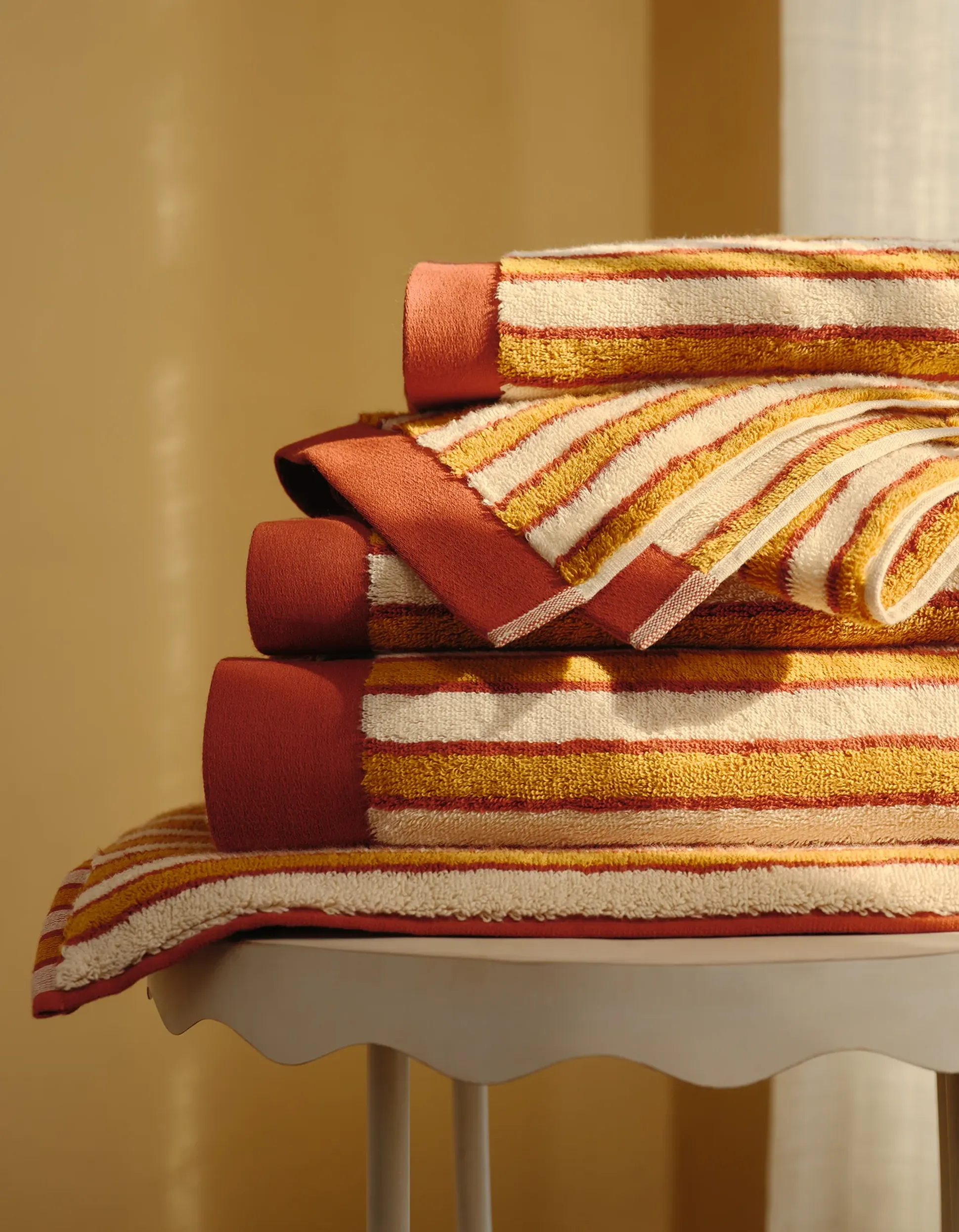 Une pile de serviettes rayées aux couleurs chaudes posée sur une petite table claire, devant un fond jaune pâle.