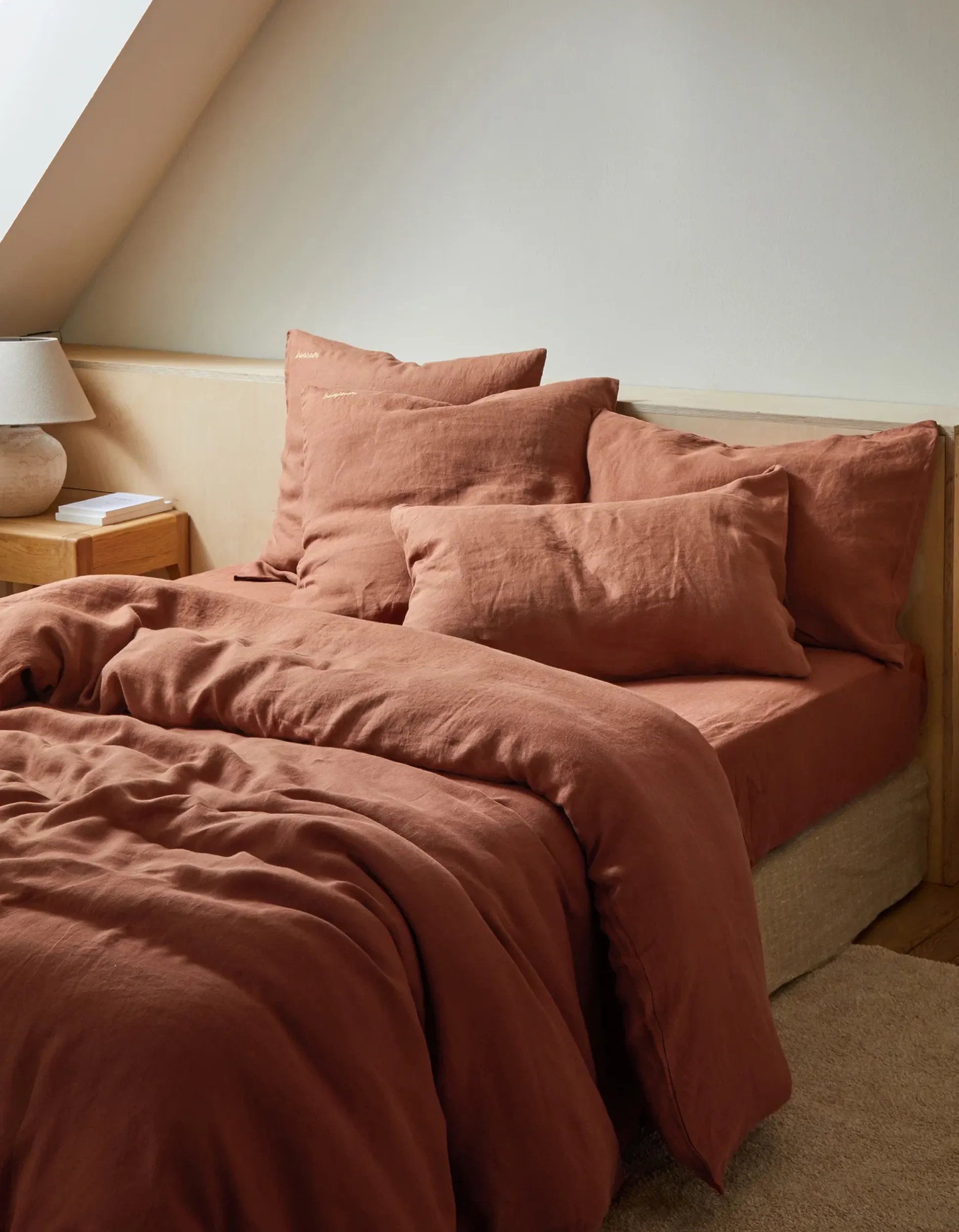 Un lit douillet recouvert d'un couvre-lit et d'oreillers couleur rouille, à côté d'une table de chevet en bois sur laquelle se trouvent une lampe et des livres, sous un plafond en pente.