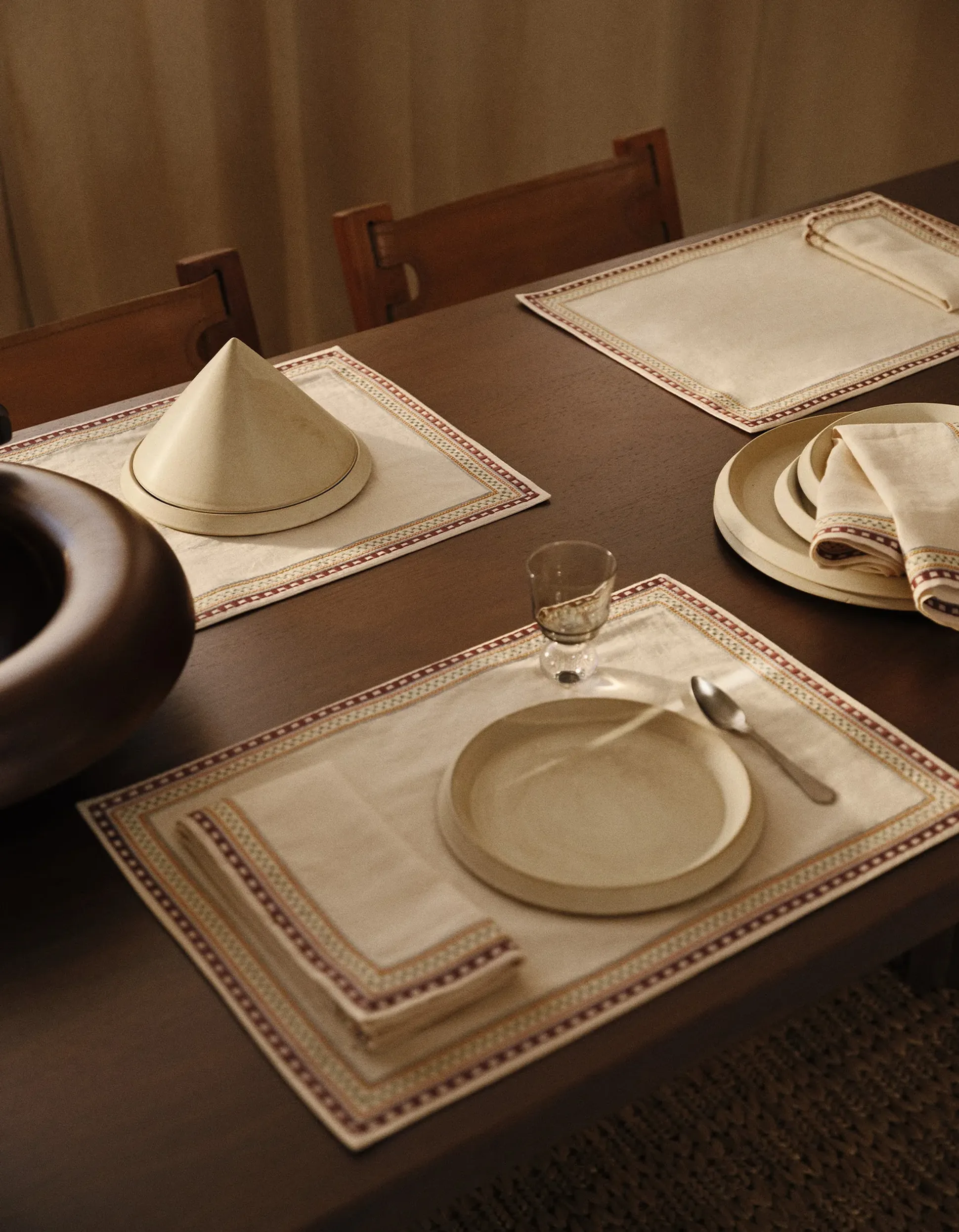Elegant dining table with beige placemats and napkins with colorful trim, featuring plates, cutlery, a glass, and a unique conical cover.