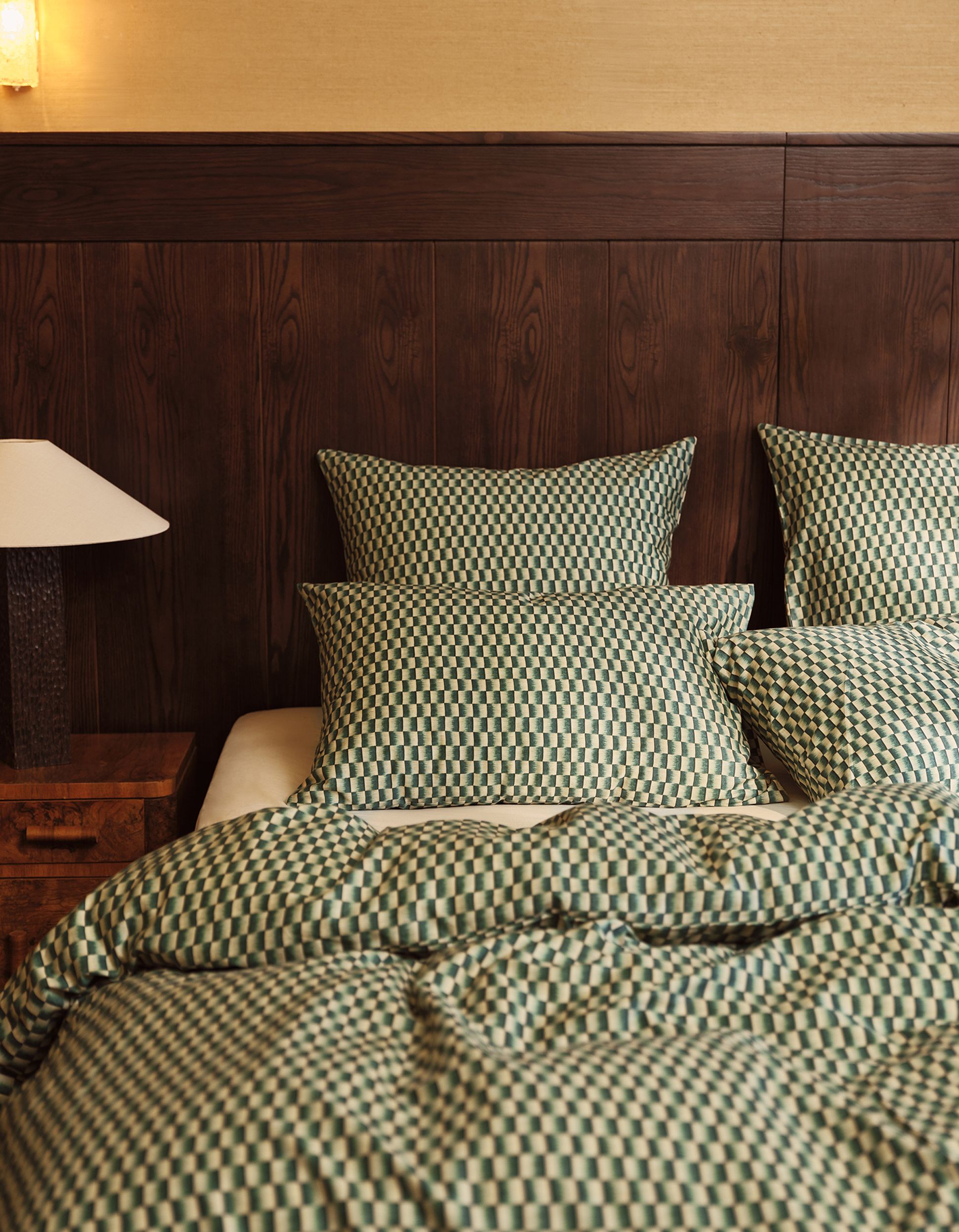 A cozy bed with checkered green bedding, three pillows, a bedside lamp, and a wooden headboard.