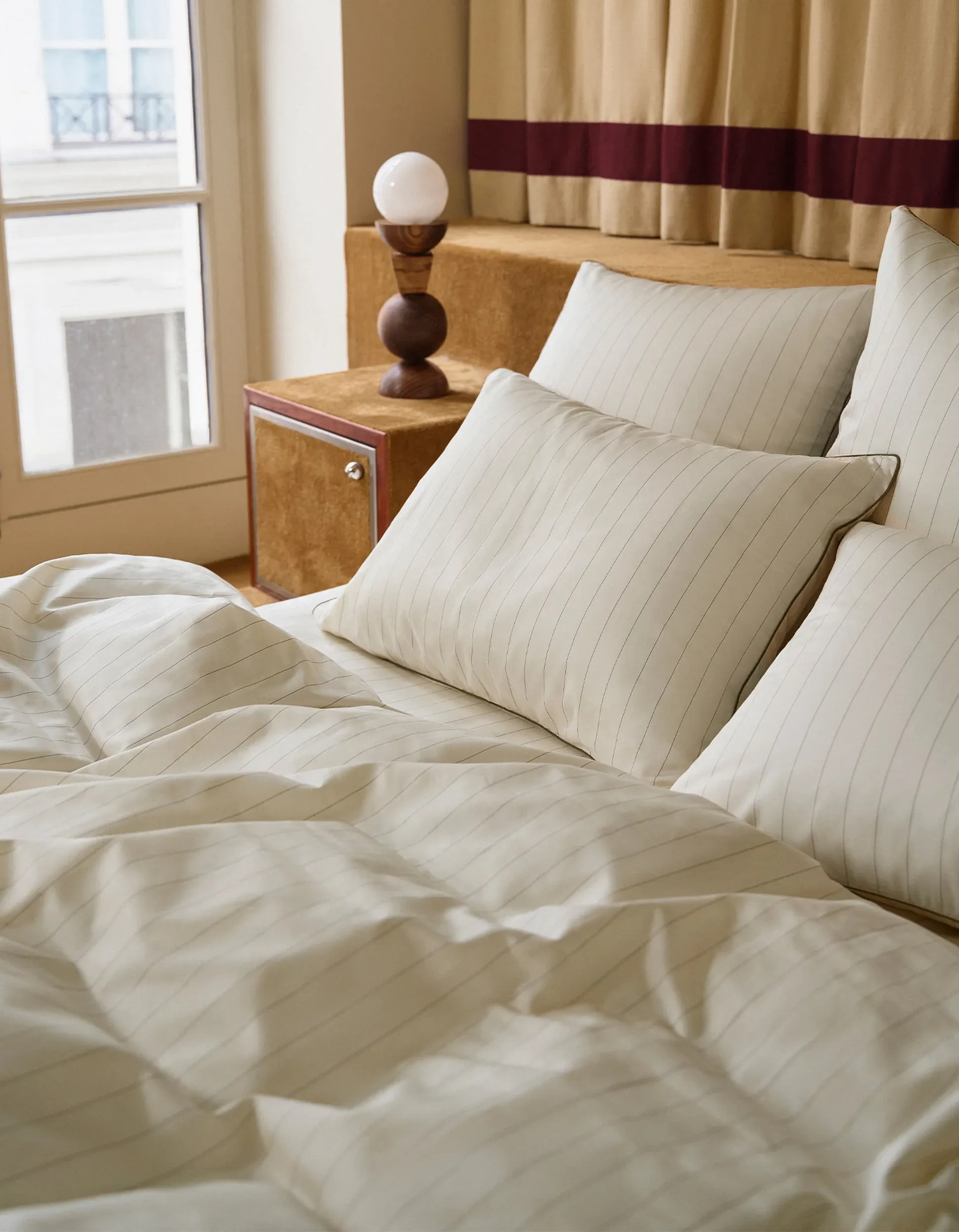 Chambre à coucher avec une parure de lit blanche à rayures, une table de chevet en bois et une lampe originale en bois équipée d'une ampoule blanche ronde. Des rideaux beiges en arrière-plan.