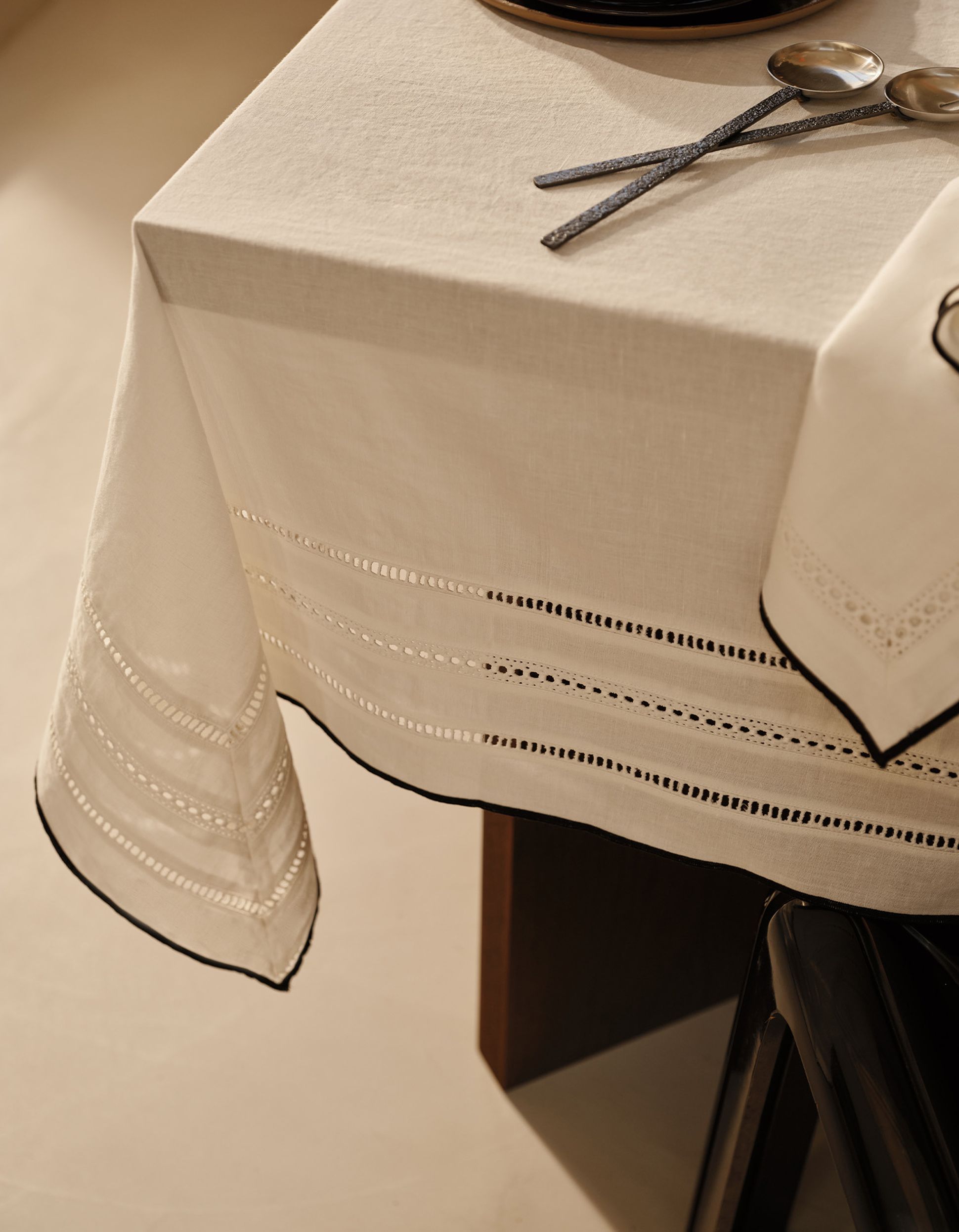 Une table soigneusement dressée, recouverte d'une nappe blanche ornée de bordures en dentelle, avec deux cuillères en argent, le tout sur un fond beige discret.