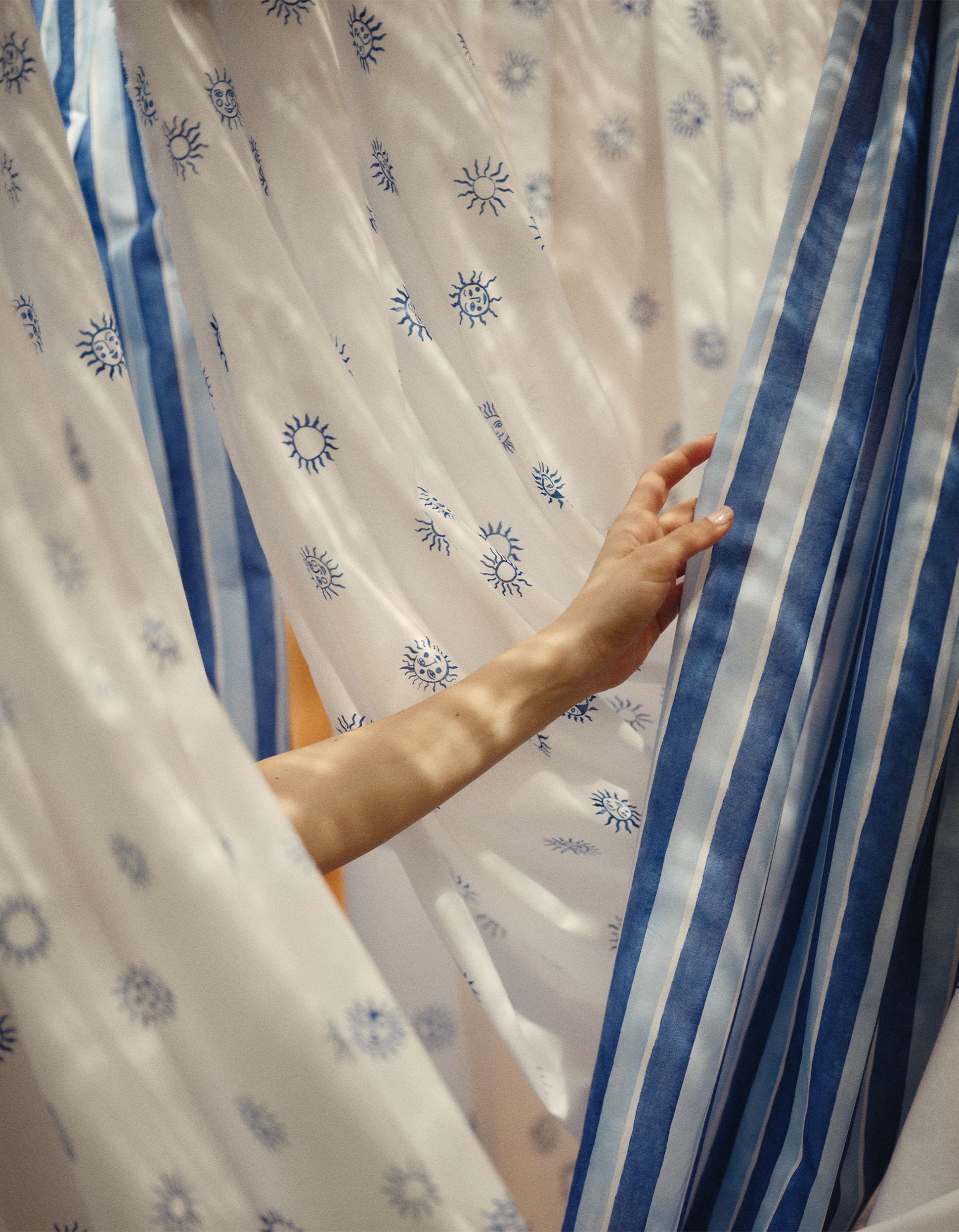 Une main écarte délicatement les rideaux à rayures bleues et à motifs solaires blancs, créant ainsi une atmosphère sereine et douce.