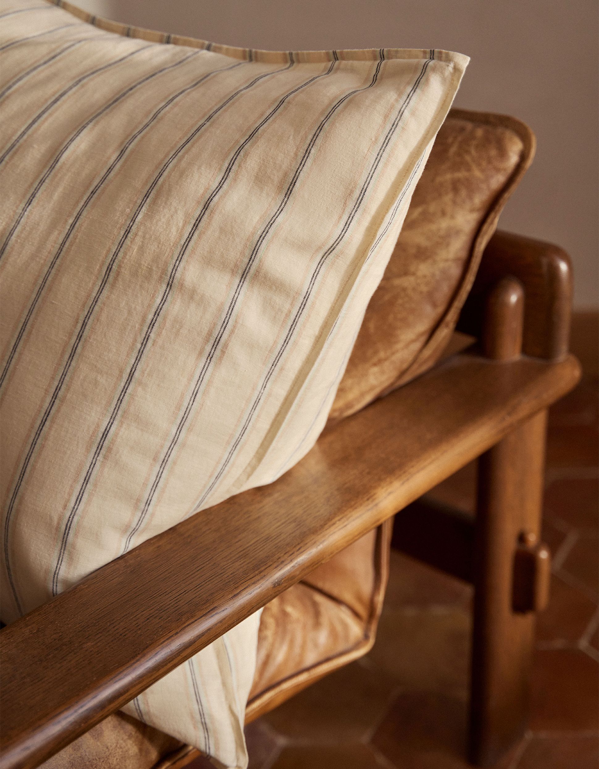 Un fauteuil en bois avec des coussins en cuir marron et un grand coussin rayé, posé devant un mur de couleur neutre sur un sol carrelé de carreaux hexagonaux.