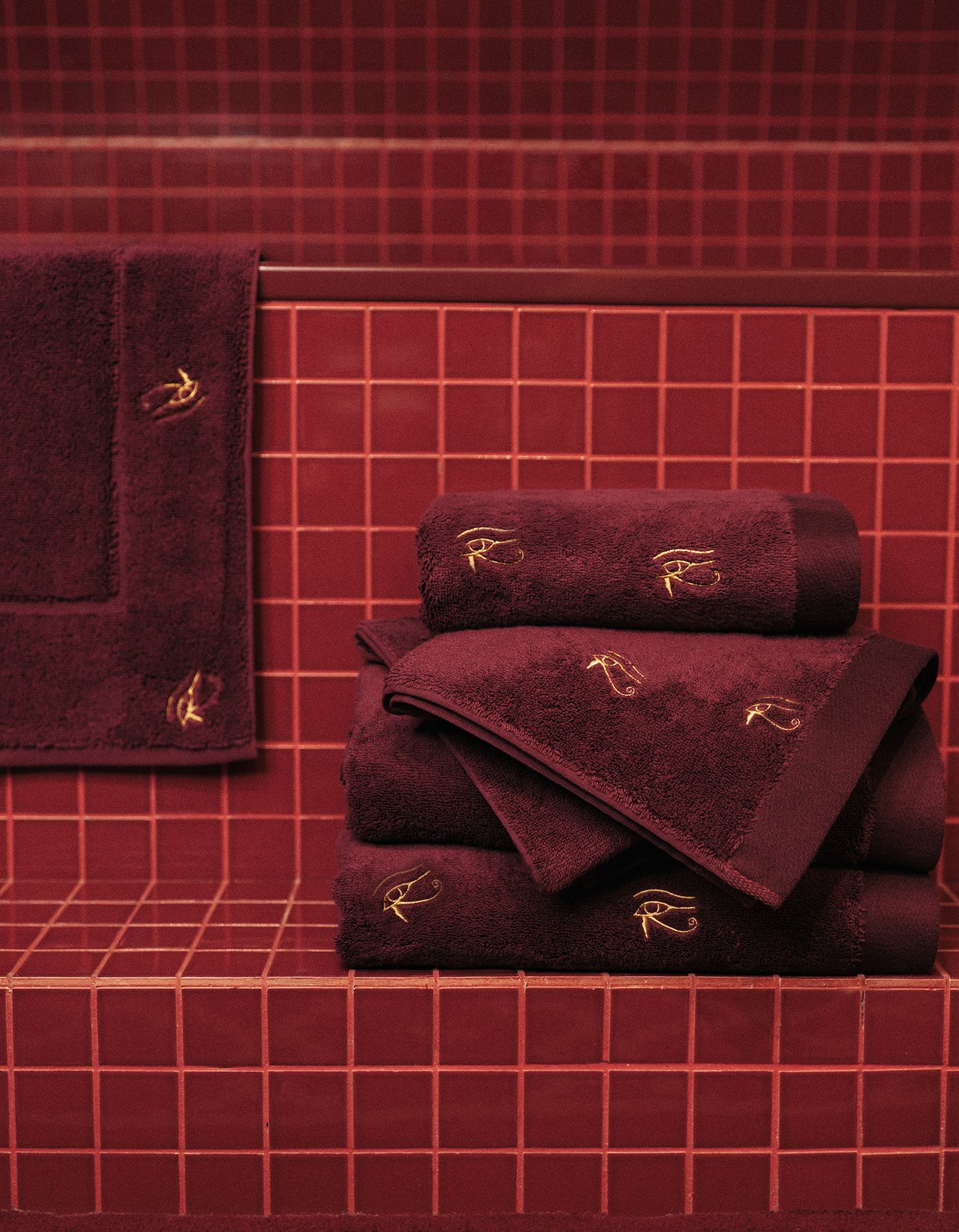 Une pile de serviettes bordeaux ornées d'yeux brodés en doré, posée sur un carrelage bordeaux, avec un mur et un tapis de bain assortis en arrière-plan.