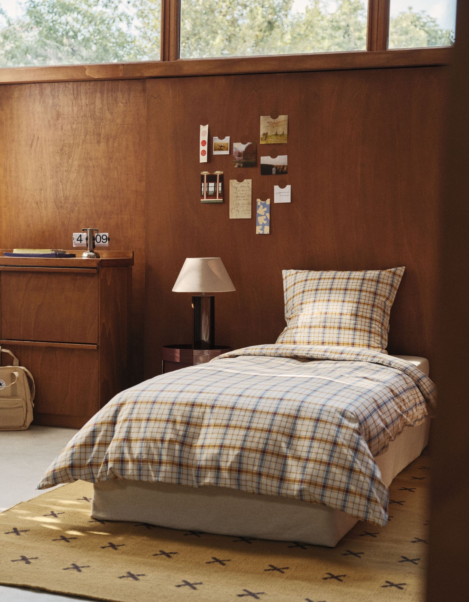 Une chambre douillette avec un lit simple recouvert d'un linge de lit à carreaux, un mur en bois orné de photos, une petite lampe et une fenêtre donnant sur la verdure à l'extérieur.