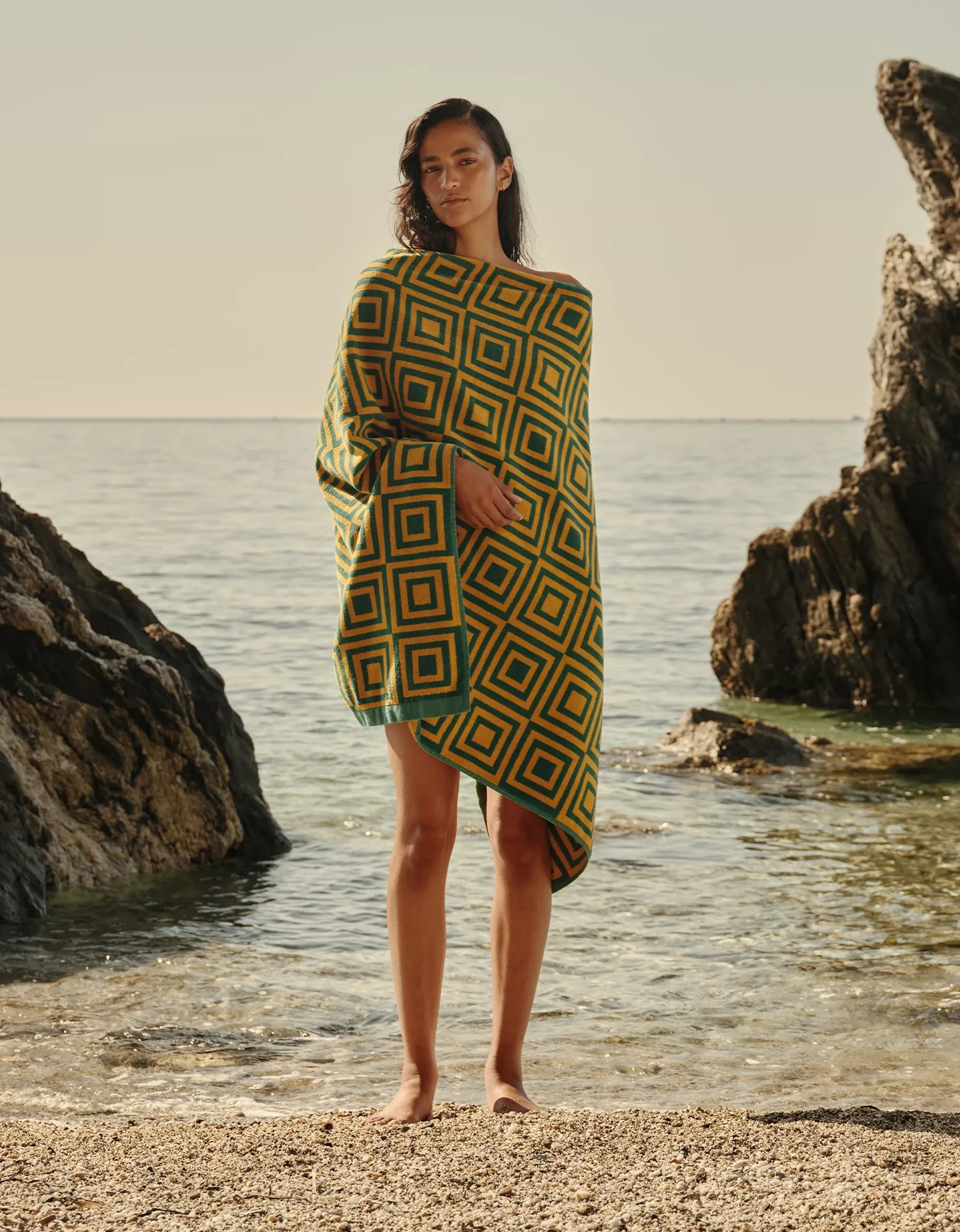 Une personne se tient debout sur une plage rocheuse, enveloppée dans un châle aux motifs géométriques dans des tons de jaune et de vert, avec l'océan serein en toile de fond.