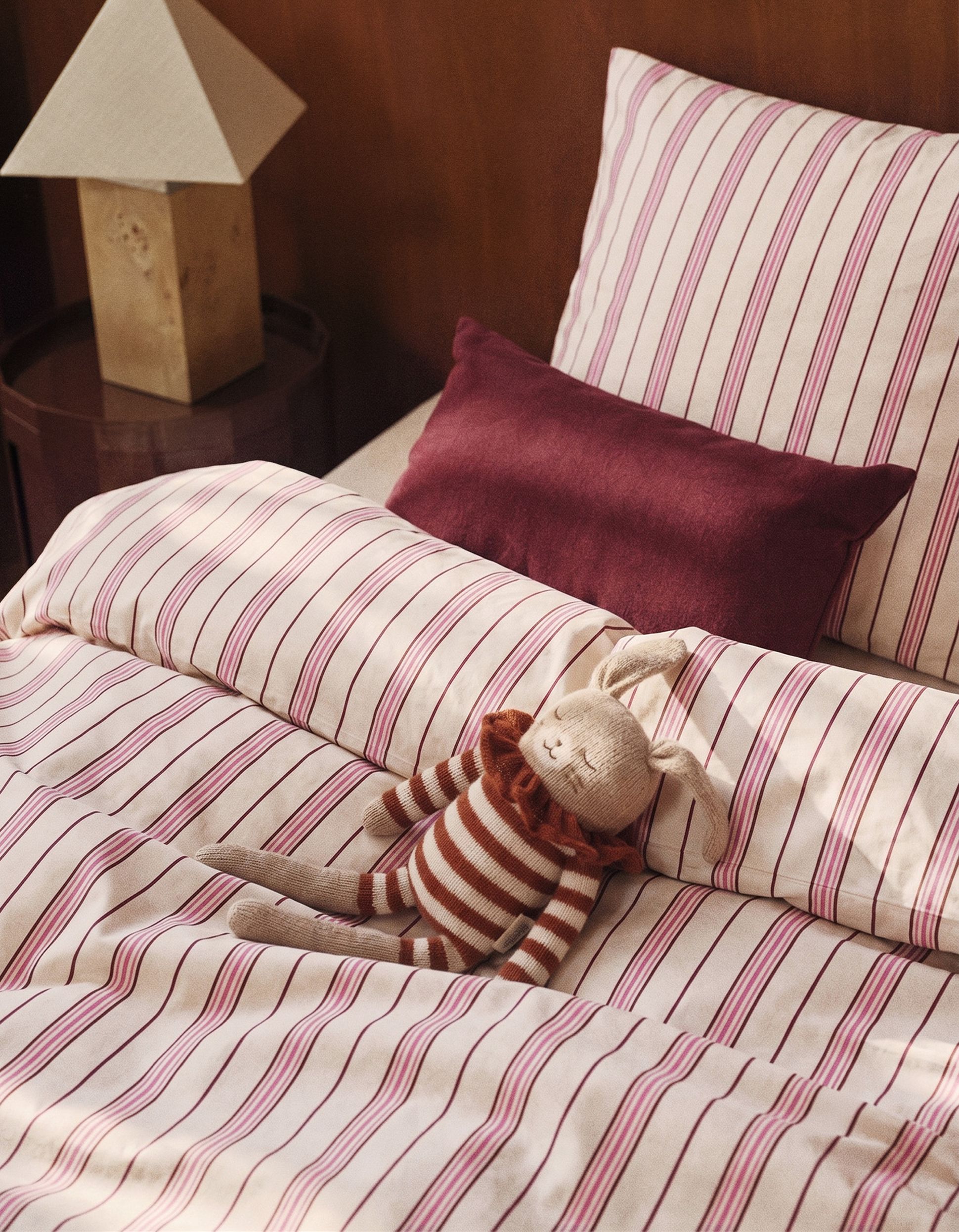 Une parure de lit rayée accompagnée d'une petite peluche en forme de lapin rayé. À l'arrière-plan, on aperçoit un coussin bordeaux et une table d'appoint sur laquelle trône une lampe moderne.