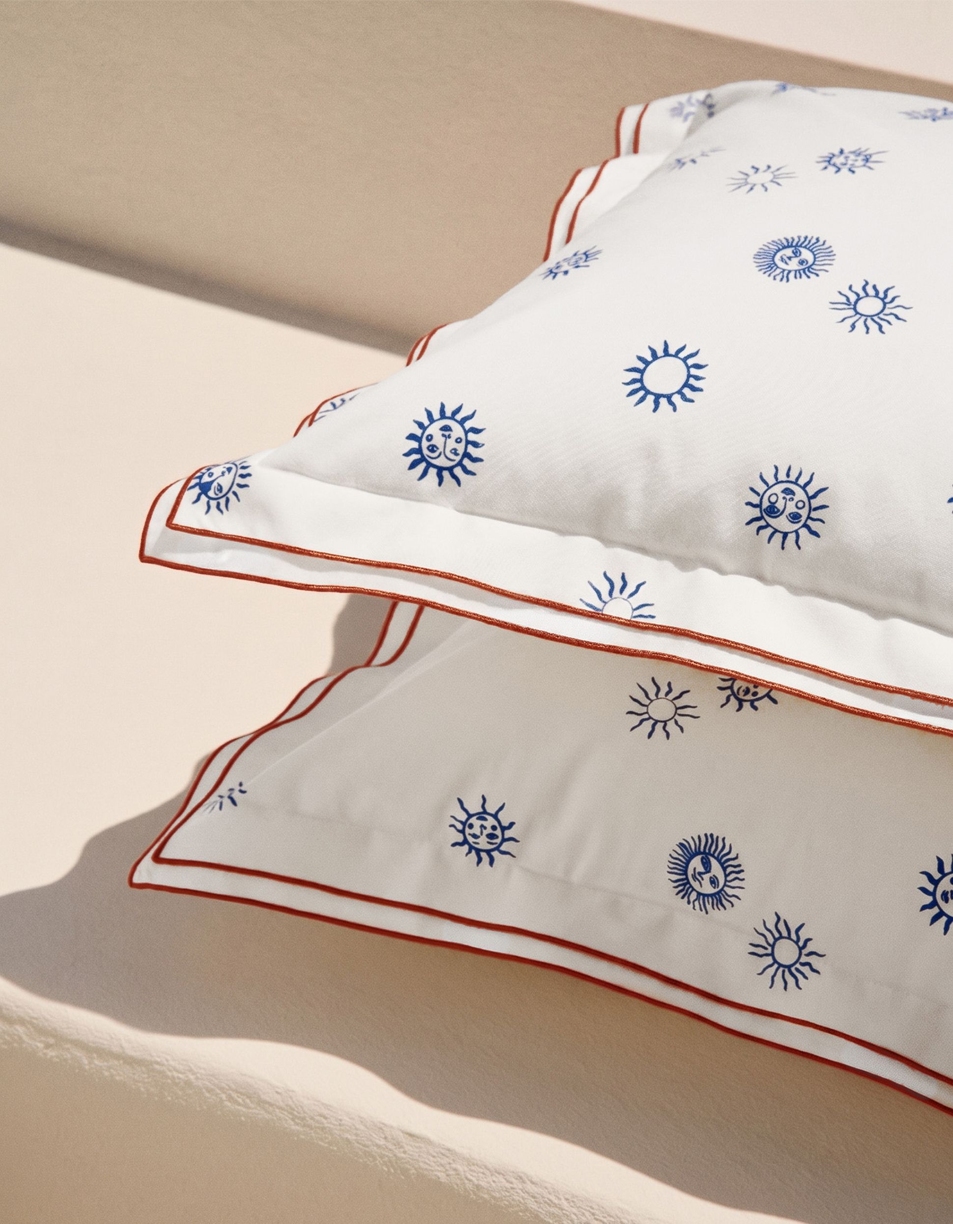 Deux coussins blancs ornés de motifs de soleils bleus et bordés de rouge sont empilés sur une surface beige.