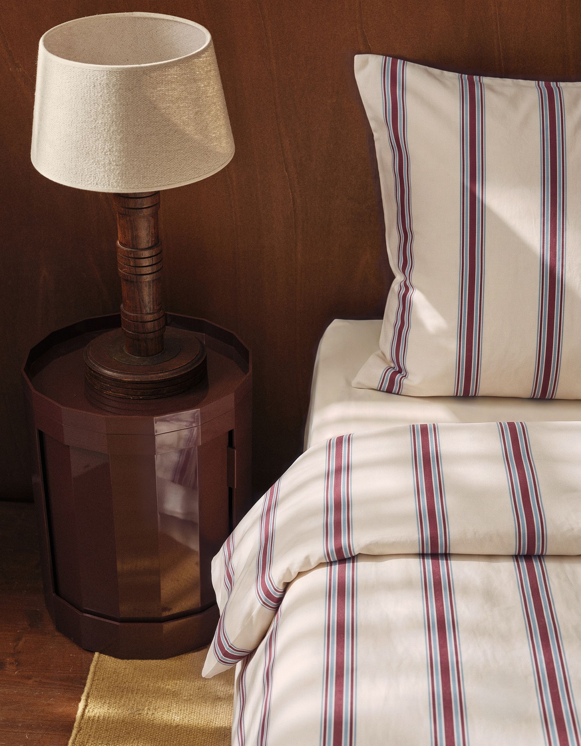 Une table de chevet sur laquelle repose une lampe marron, à côté d'un lit recouvert d'une parure de lit crème à rayures, devant un mur en bois.