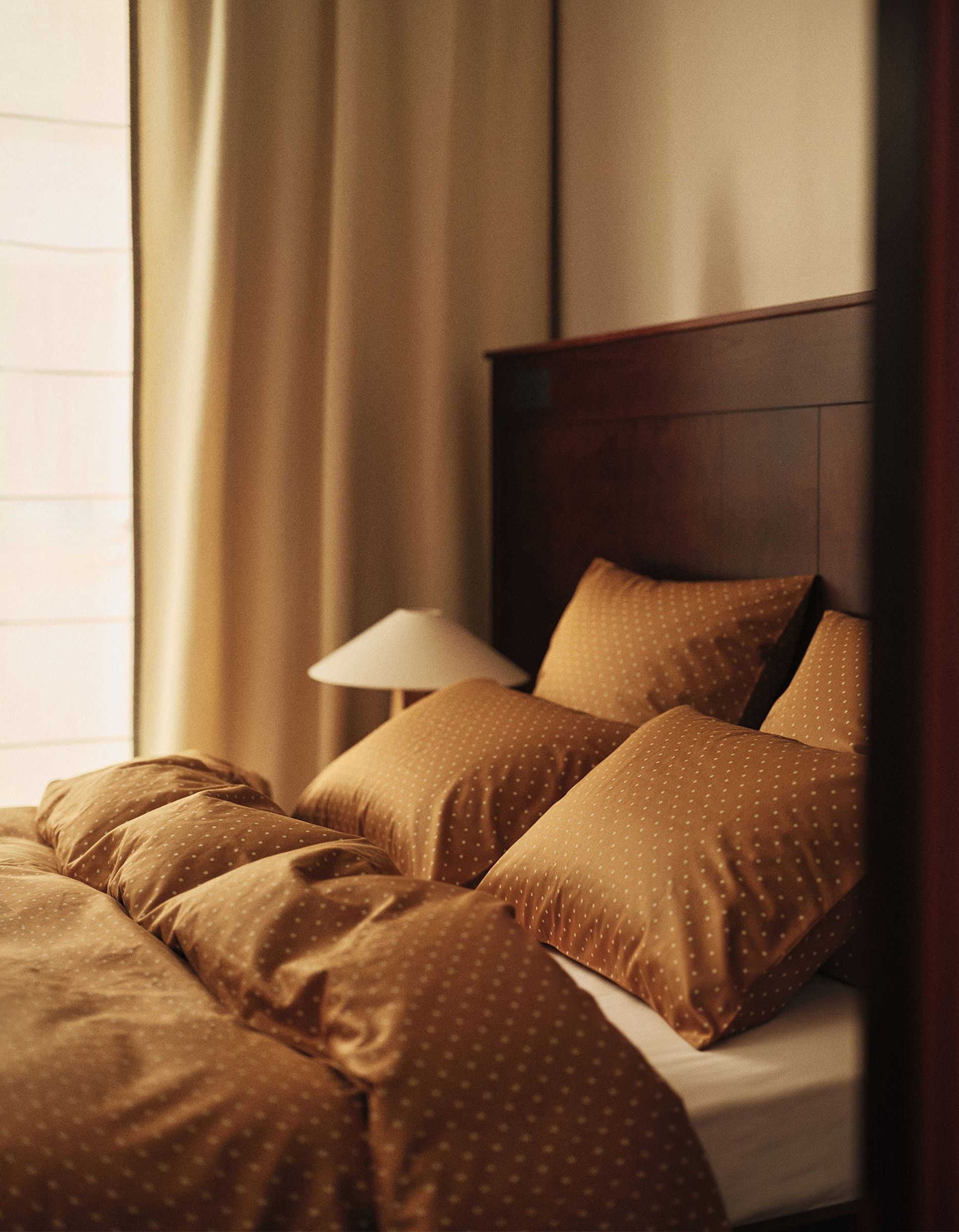 Lit doté de draps et d’oreillers à pois marron, placé à côté d’une table de chevet en bois surmontée d’une lampe, le tout adossé à une tête de lit en bois foncé.