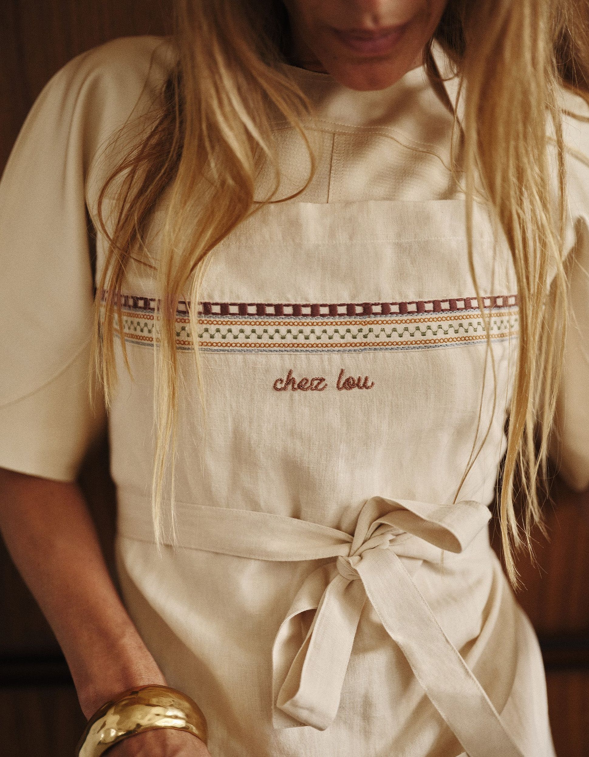 Une femme portant un tablier beige orné de motifs brodés et de l'inscription « chez lou », ses longs cheveux blonds tombant sur ses épaules.