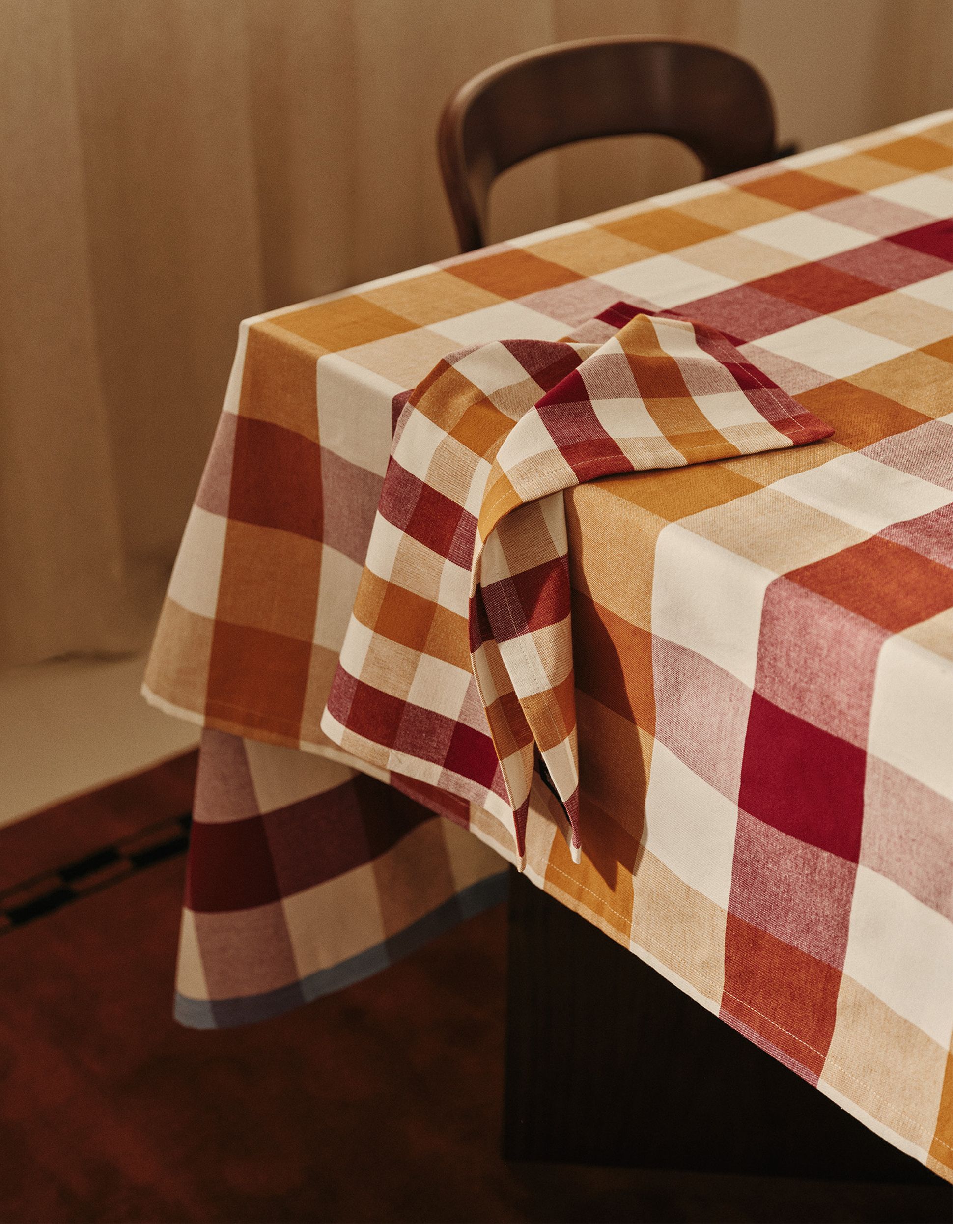 Une table recouverte d'une nappe à carreaux dans les tons orange, rouge et blanc, avec une serviette assortie posée sur le bord, à côté d'une chaise en bois.
