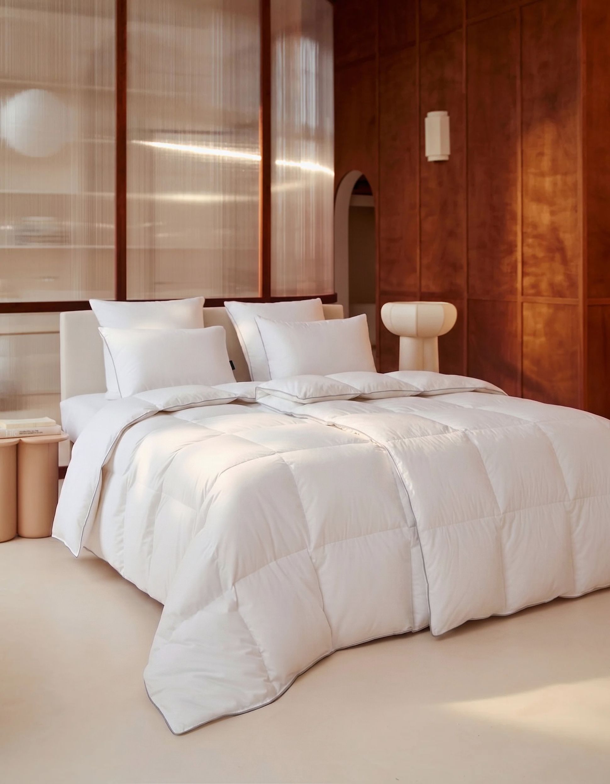 A modern bedroom with a neatly made bed, white pillows, and a soft duvet. The room features warm wooden paneling and a minimalist design.