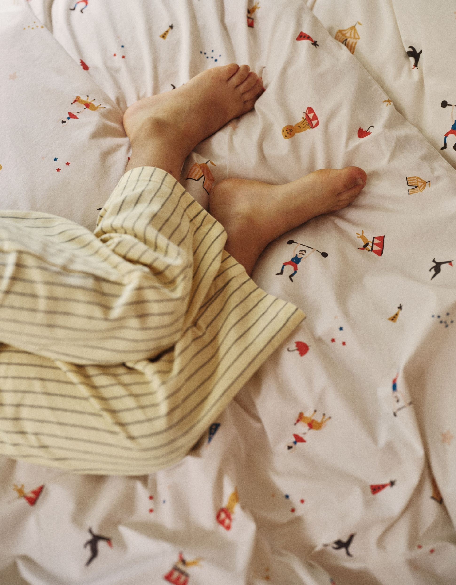 Les pieds d'un enfant, vêtu d'un pyjama rayé, reposent sur un lit orné d'un motif sur le thème du cirque.