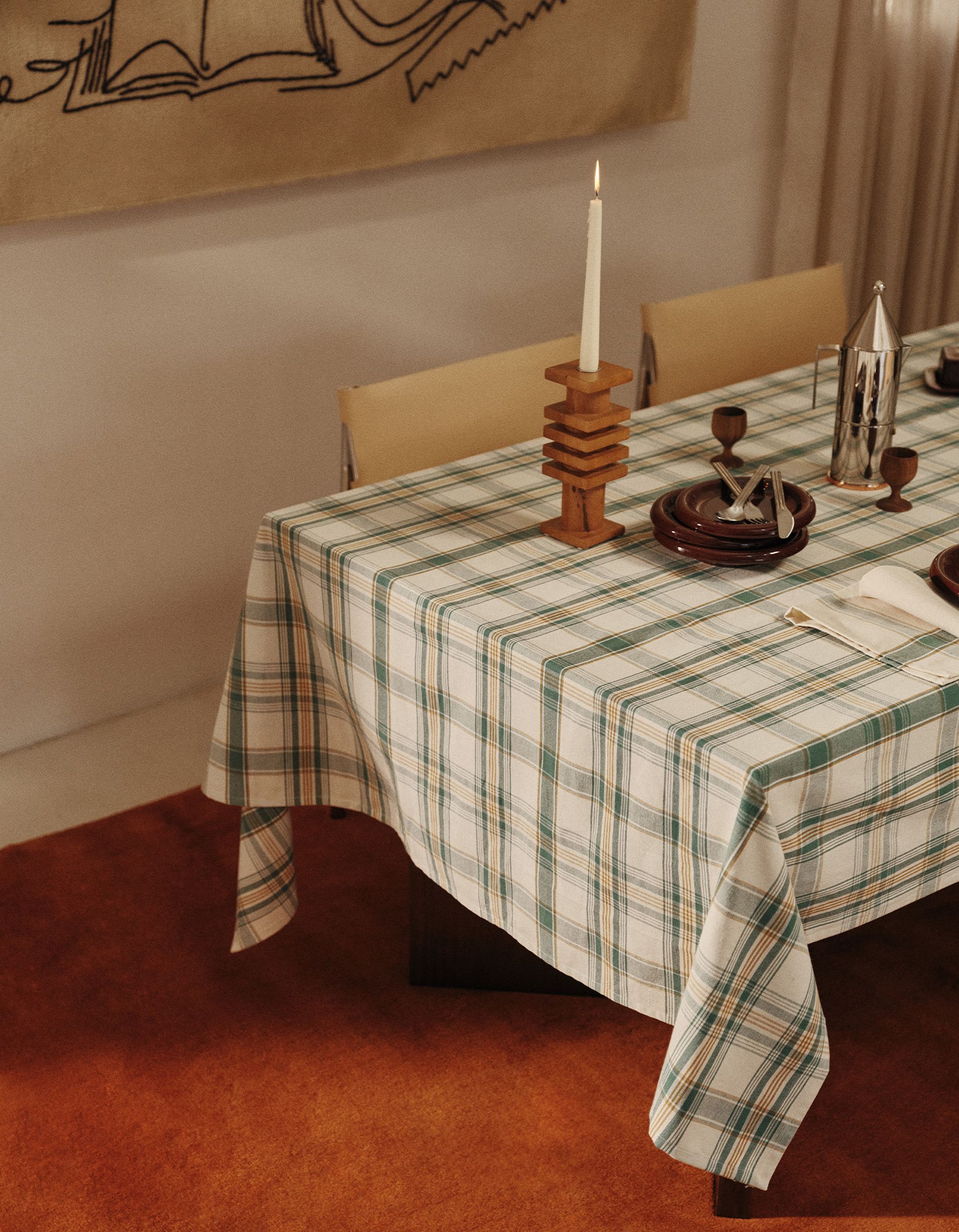 Une table recouverte d'une nappe à carreaux, sur laquelle sont disposés un bougeoir en bois, une bougie allumée, des assiettes, des couverts, des tasses et une cafetière en métal, le tout posé sur un tapis.