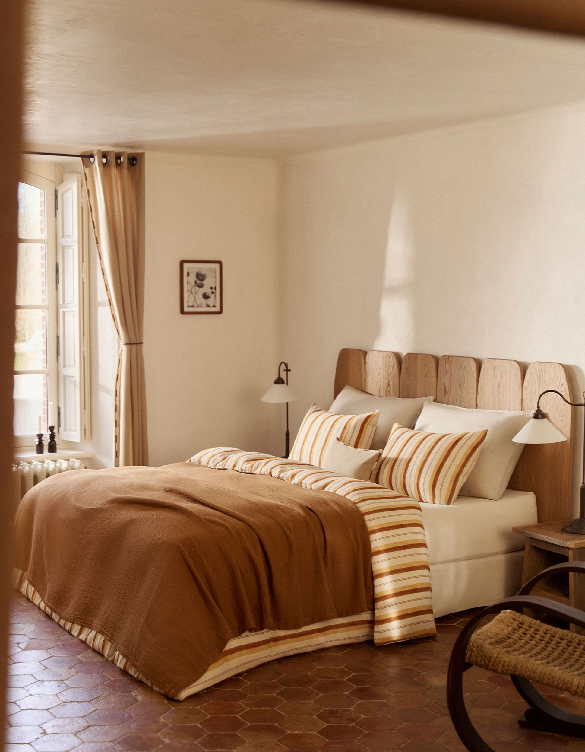 Chambre confortable avec une tête de lit en bois, une literie rayée, deux lampes, un tableau encadré, des rideaux beiges et un sol en carreaux de terre cuite.
