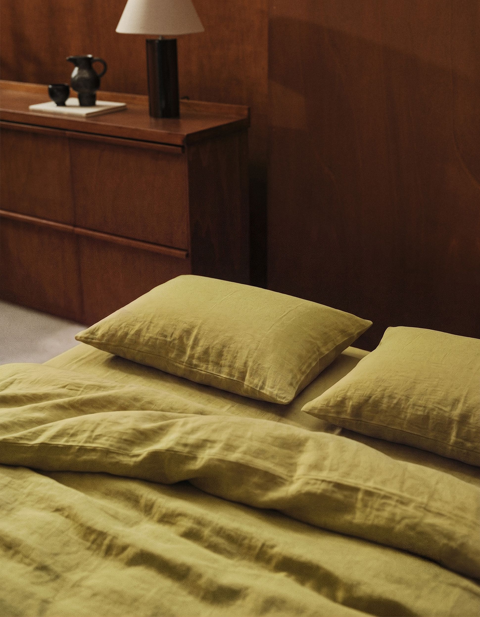 Une chambre douillette comprenant un lit avec des draps couleur moutarde et deux oreillers, une commode en bois surmontée d'une lampe noire, ainsi qu'un service à thé.
