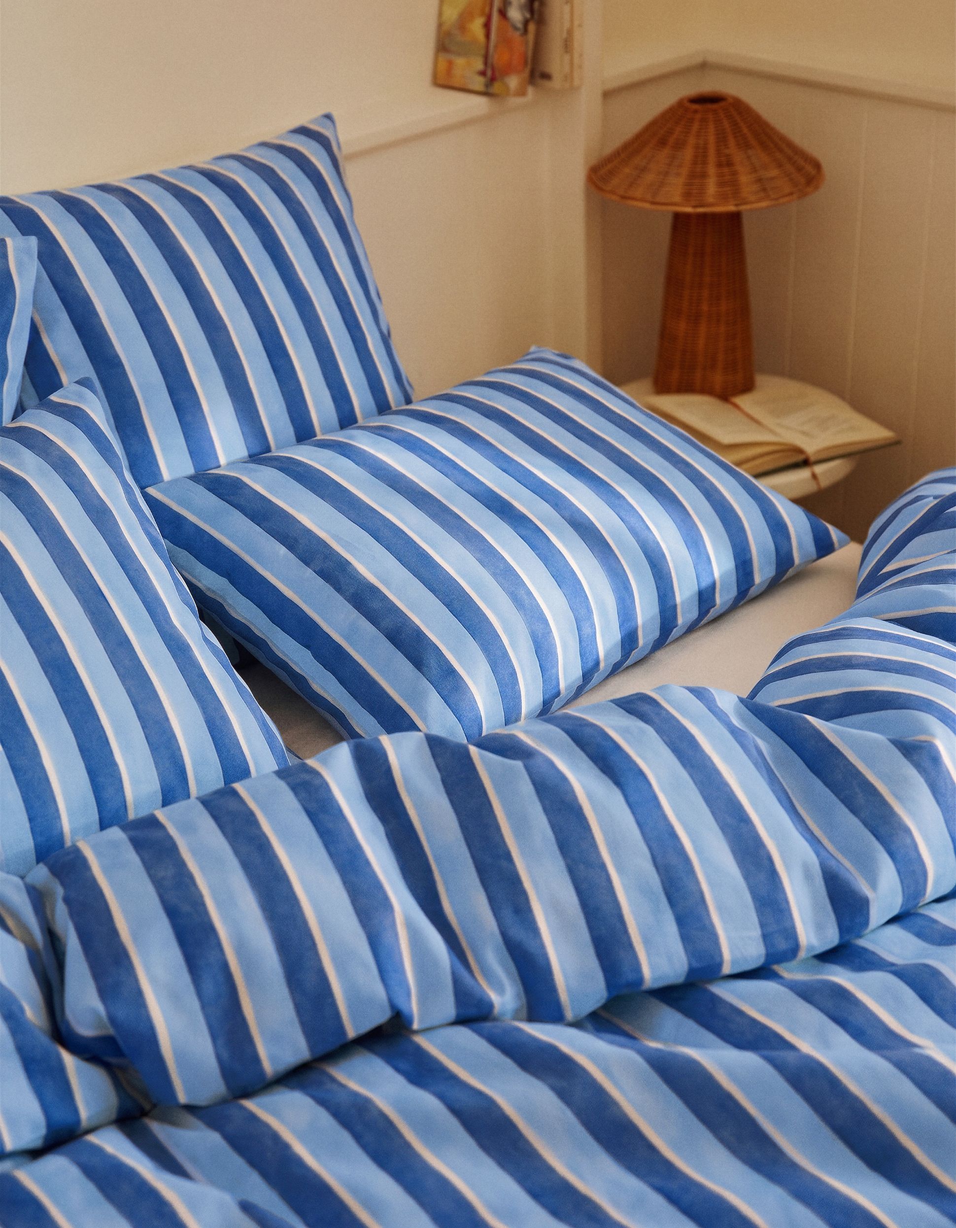 Un ensemble de literie à rayures bleues et blanches sur un lit, avec une lampe en osier et un livre ouvert sur la table de chevet, dans un coin douillet.