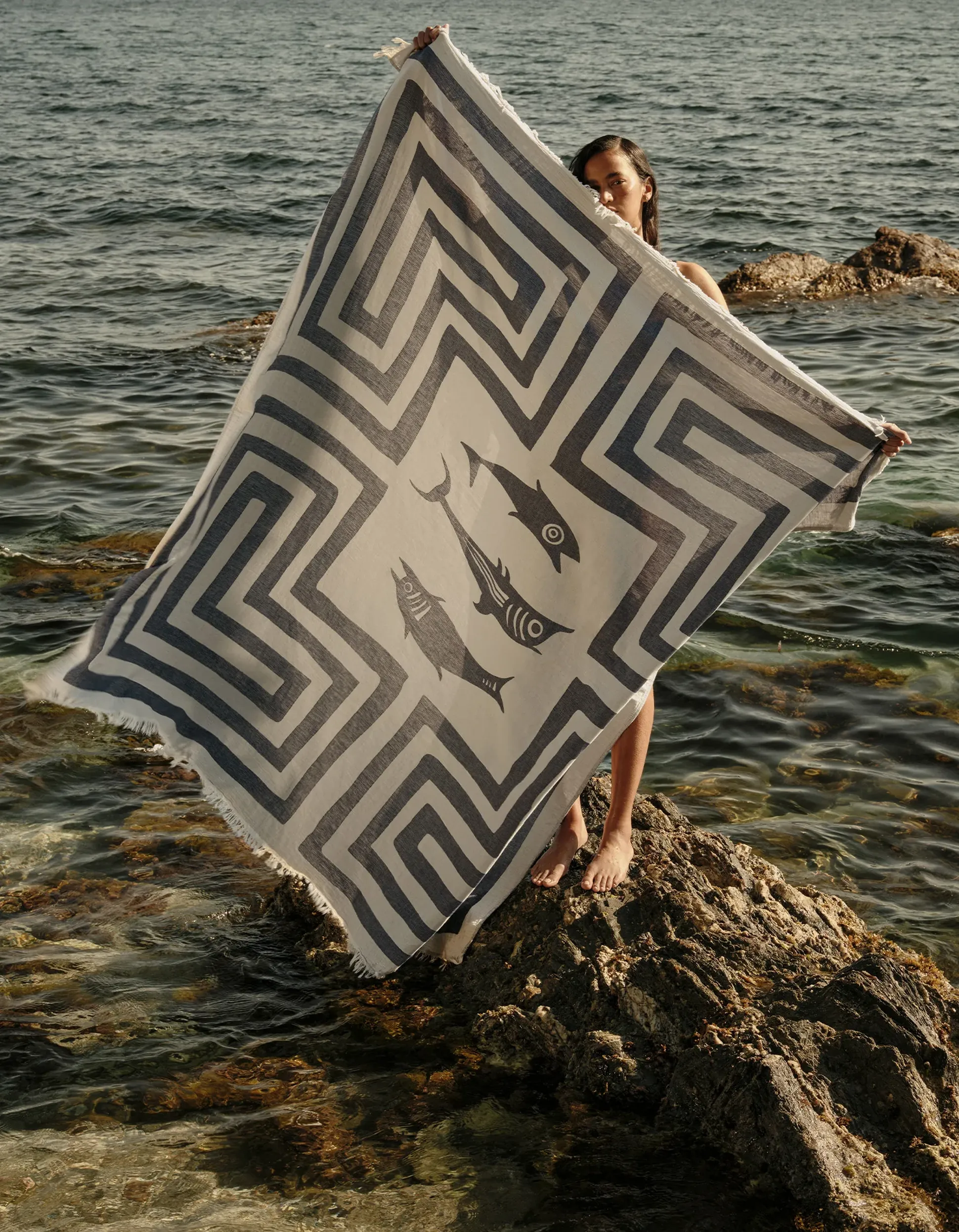 Personne tenant une grande serviette à motifs représentant des poissons, debout sur un rivage rocheux avec la mer en arrière-plan.