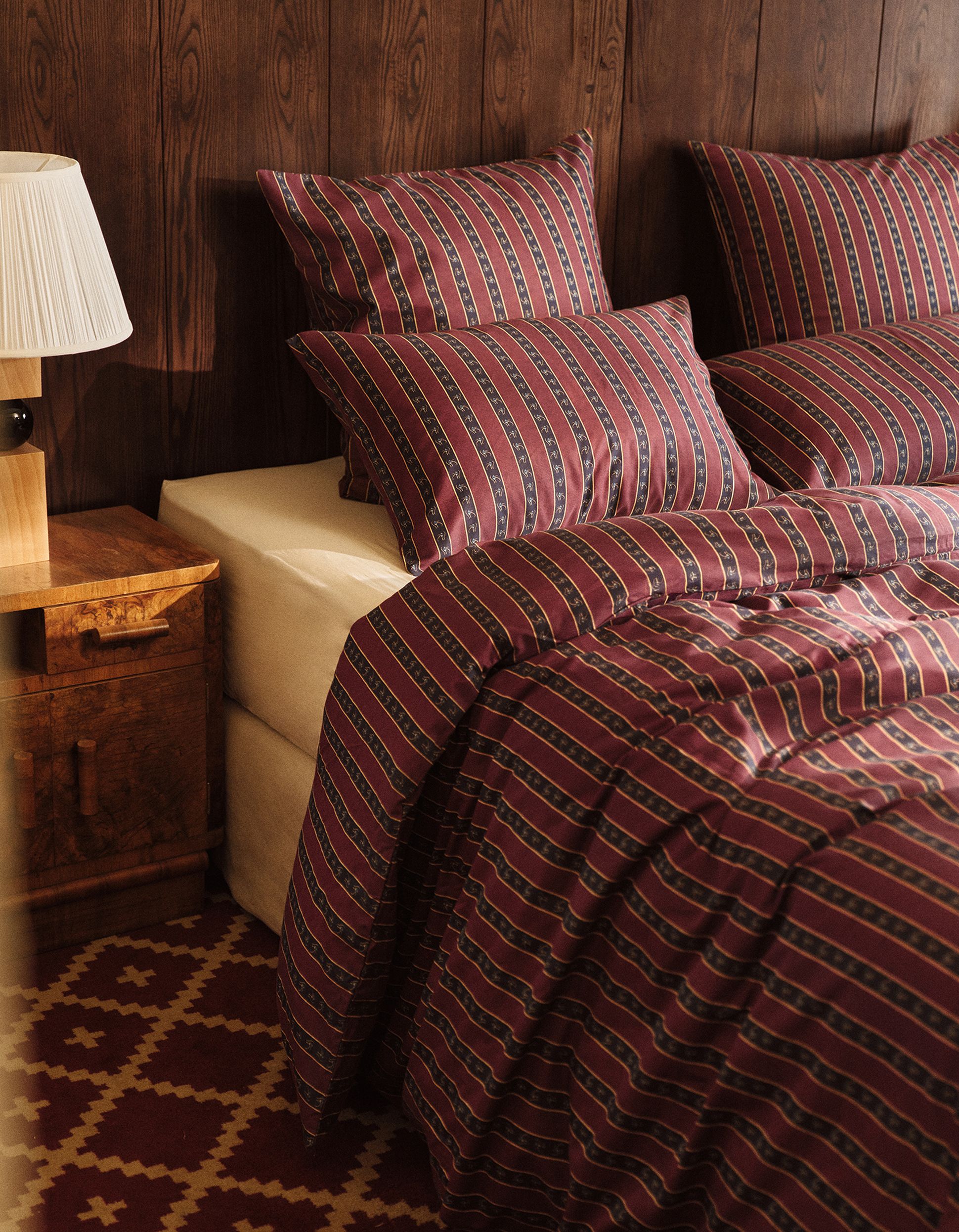 A cozy bedroom with a bed featuring striped maroon and black bedding, matching pillows, a wooden nightstand with a lamp, and a geometric patterned rug.
