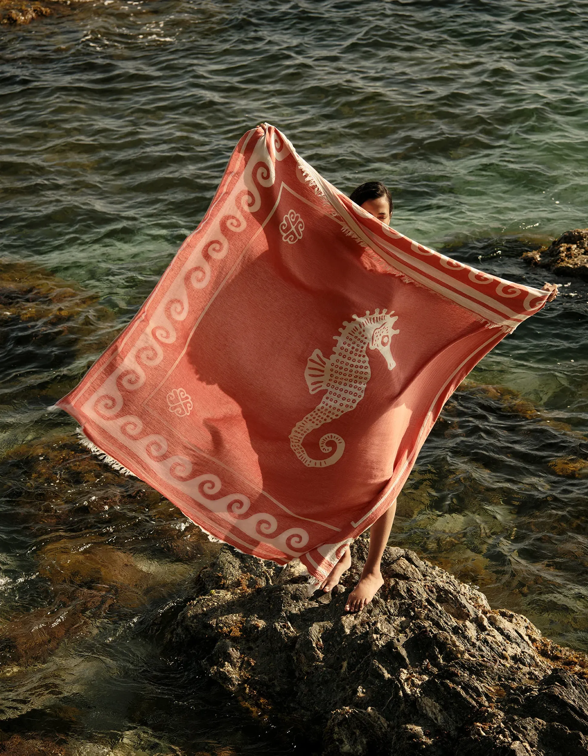 Une femme se tient debout sur un rivage rocheux, enveloppée dans une serviette rouge et blanche ornée d'un motif d'hippocampe, avec la mer en arrière-plan.
