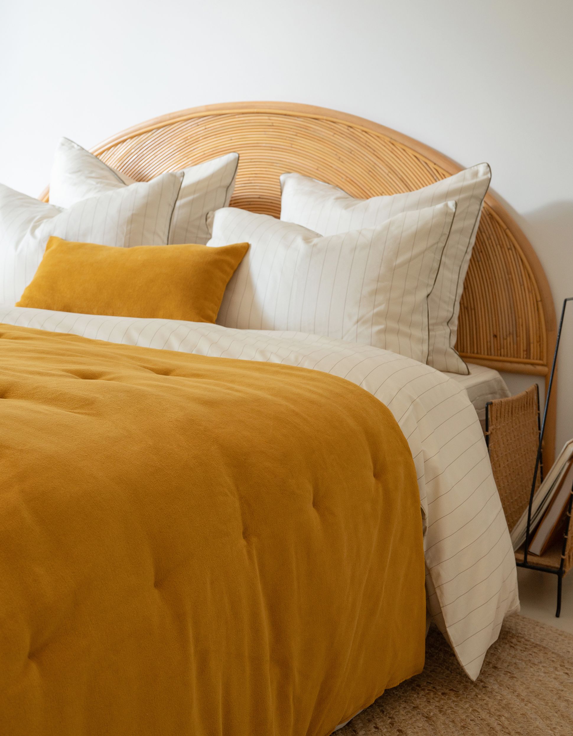 Une chambre douillette avec une tête de lit en rotin, une couverture couleur moutarde et des coussins blancs à rayures, créant une atmosphère chaleureuse et accueillante.
