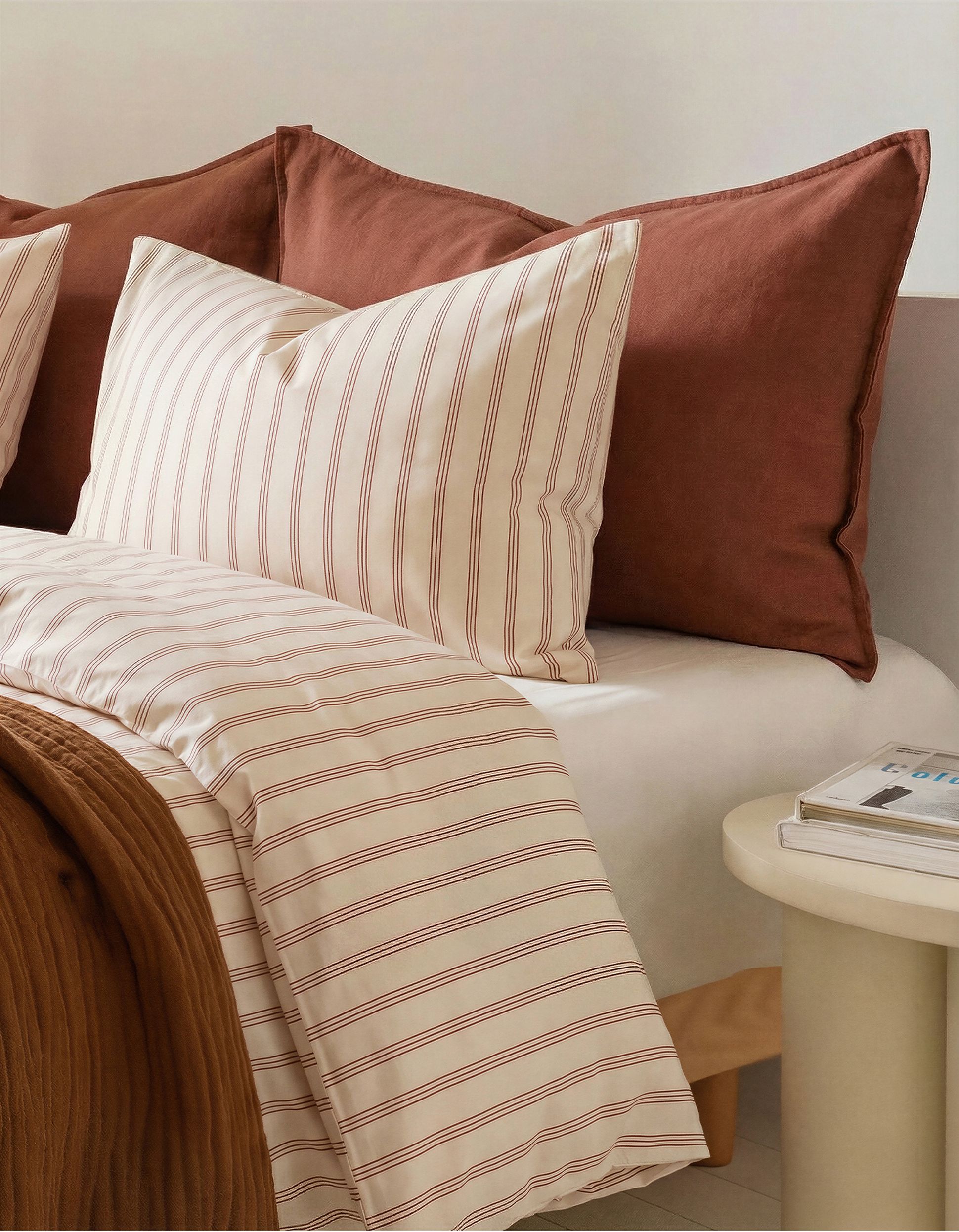 Lit douillet avec des oreillers rayés et une couette assortie dans des tons neutres, complété par une couverture marron et une petite table d'appoint blanche adjacente.