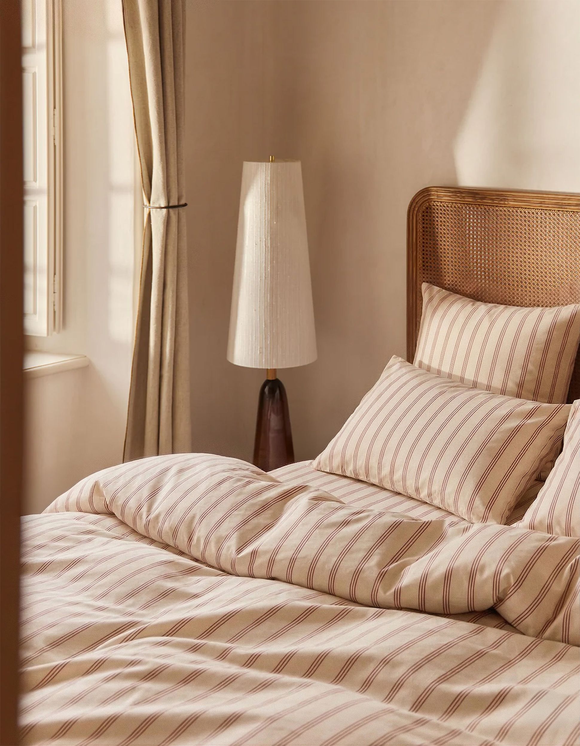 Une chambre douillette avec une parure de lit rayée, une tête de lit en rotin, un lampadaire et des rideaux légers près d'une fenêtre baignée de soleil, créant une atmosphère chaleureuse et accueillante.