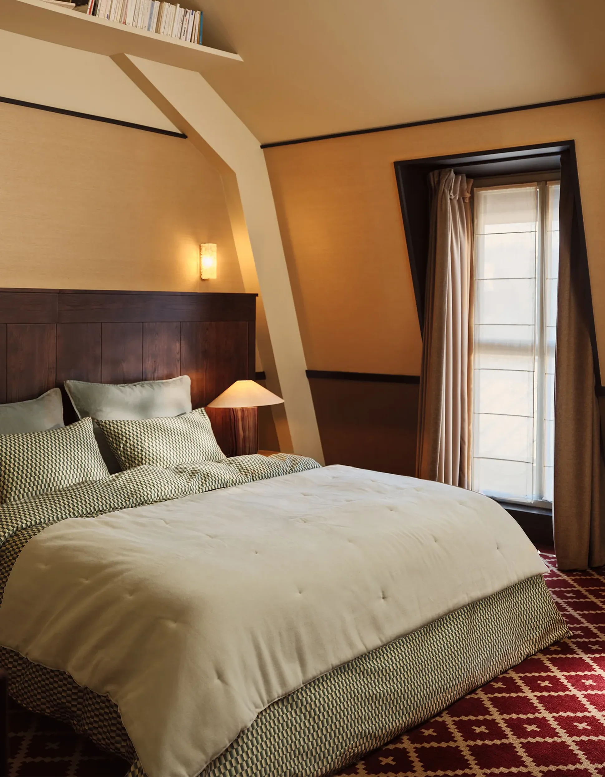 Chambre à coucher chaleureuse avec un grand lit, une parure de lit à motifs, un éclairage tamisé, une tête de lit en bois, des murs texturés et une fenêtre triangulaire ornée de rideaux.