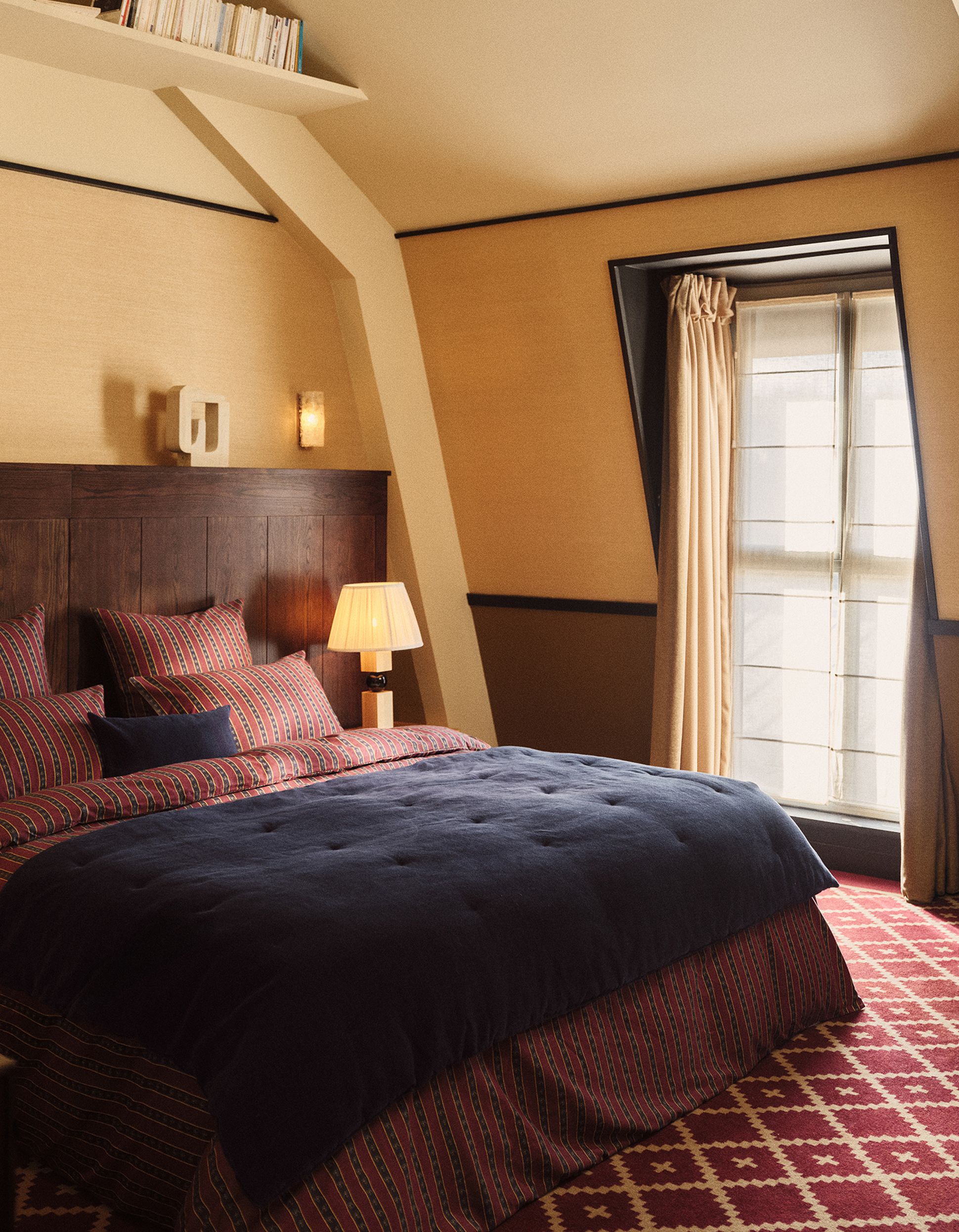 Cozy bedroom with a double bed, dark blue comforter, patterned bedspread, wooden headboard, and a bedside lamp near a window with curtains.