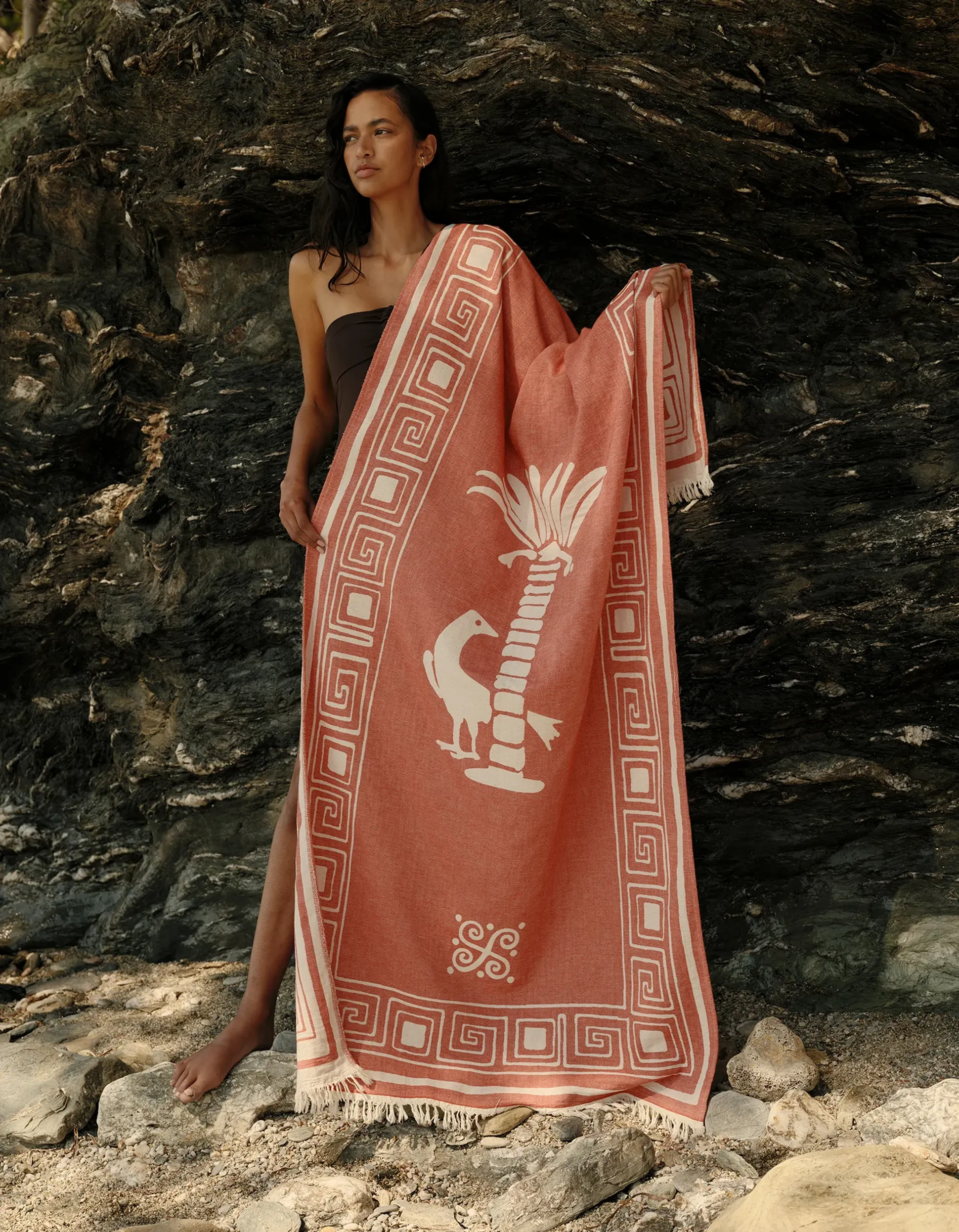 Une femme se tient debout devant un décor rocheux, tenant dans ses mains un tissu à motifs rouges sur lequel sont imprimés un oiseau et un palmier blancs ; elle porte une robe sans bretelles.