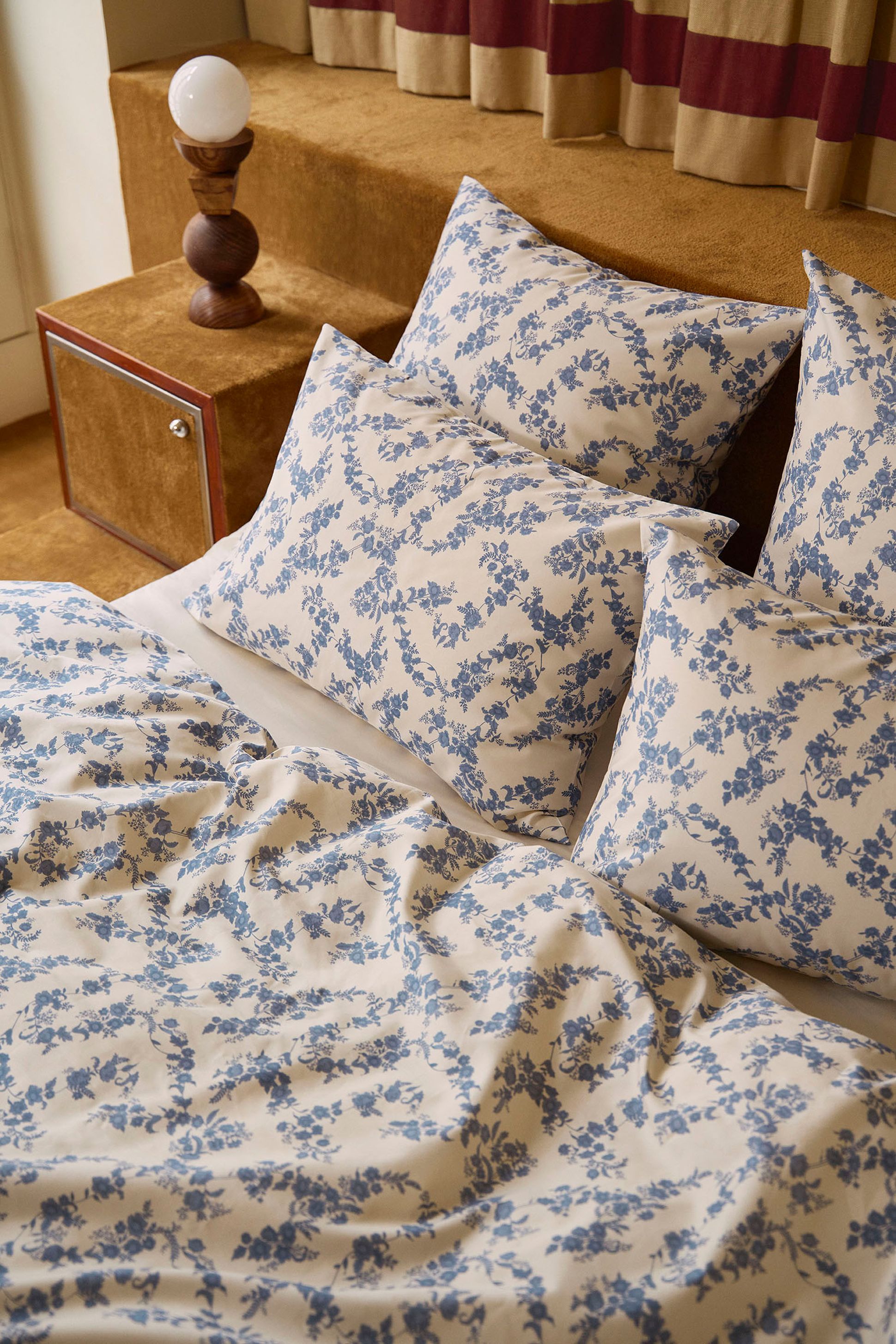 Un lit douillet avec une literie à motifs floraux bleus, une table d'appoint en bois, une lampe ronde et des rideaux beiges en arrière-plan.