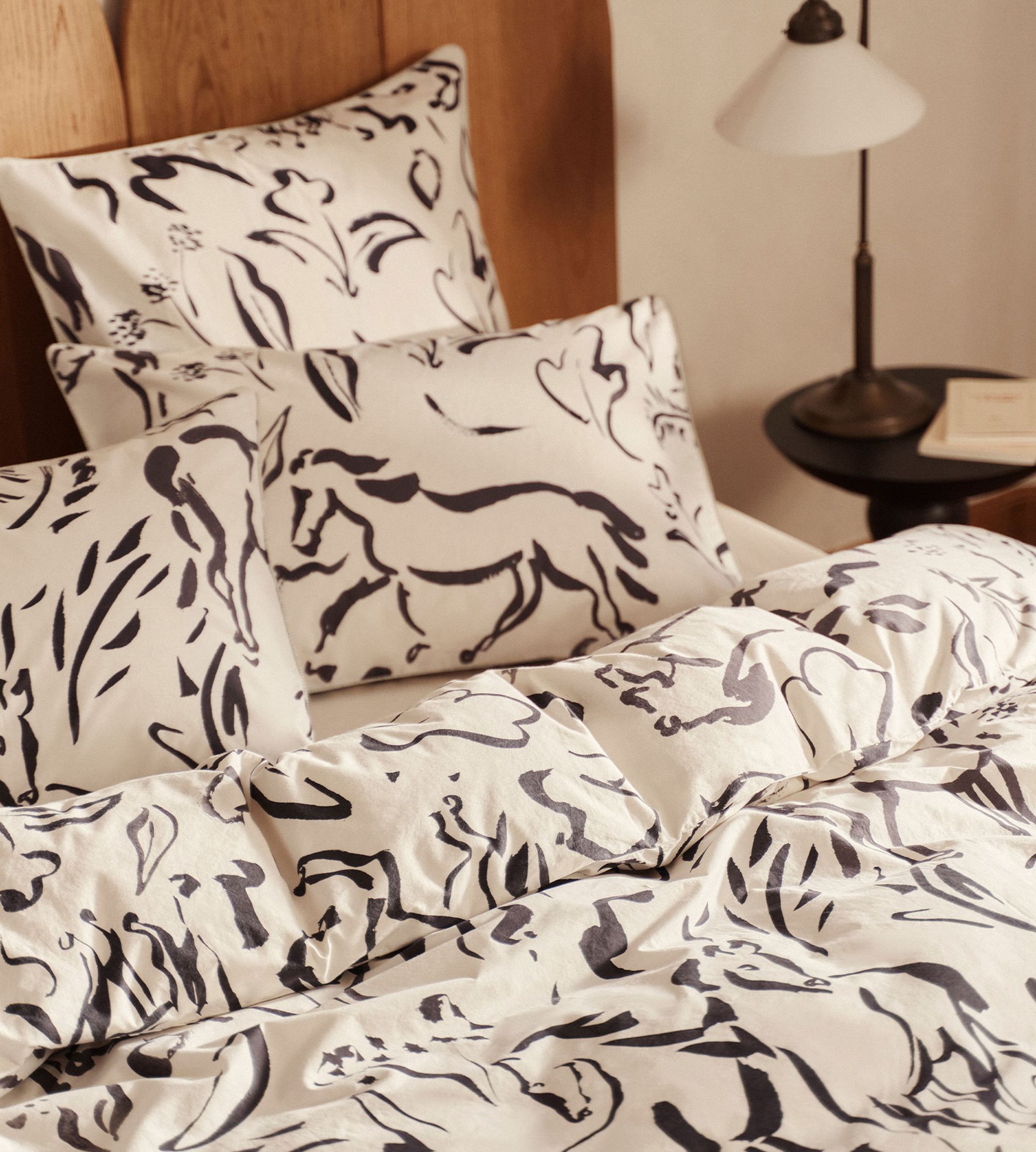 Ensemble de literie avec motifs abstraits de chevaux noirs sur des draps et des taies d'oreiller blancs, avec une tête de lit en bois et une table de chevet.