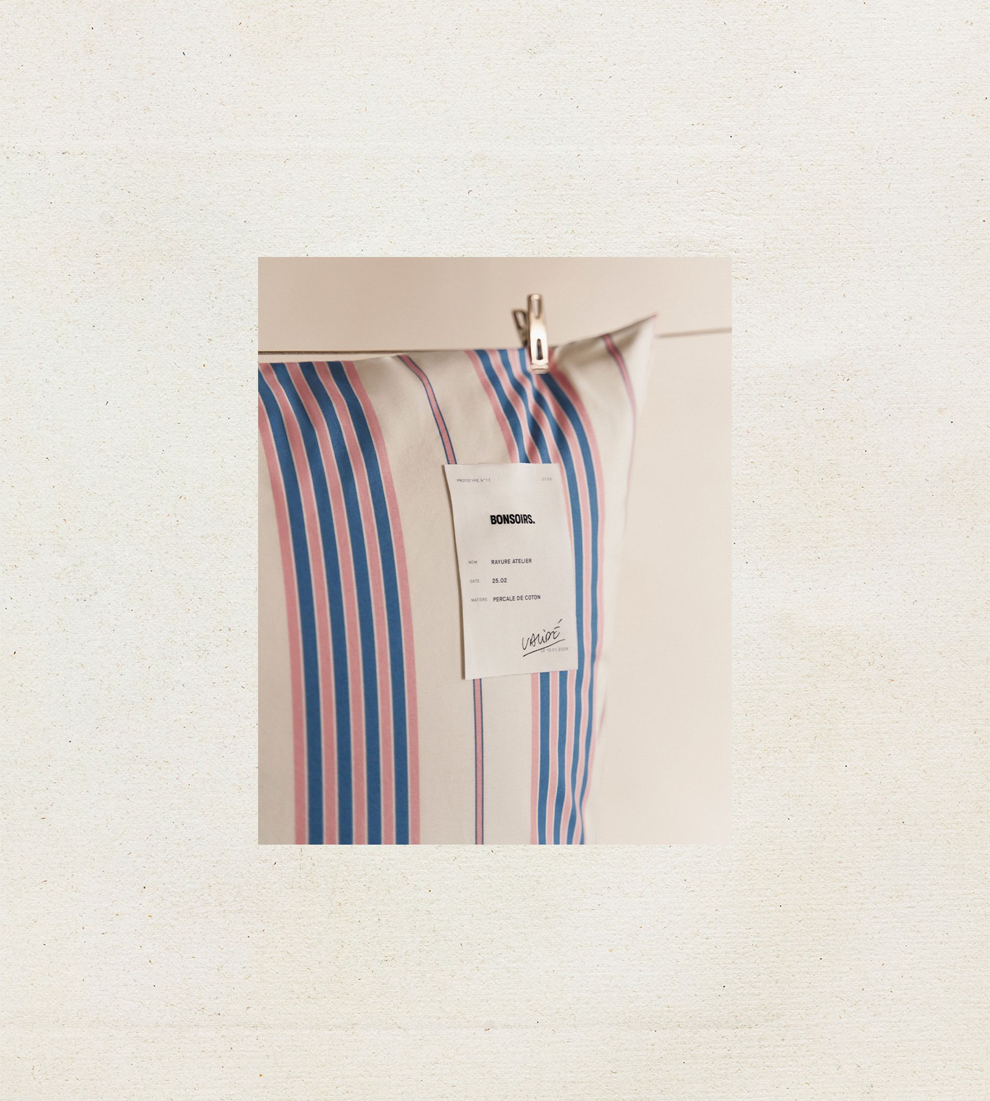 Un oreiller à rayures bleues et roses, orné d'une étiquette blanche portant l'inscription « BONSOIRS », est suspendu à un clip sur un fond clair.