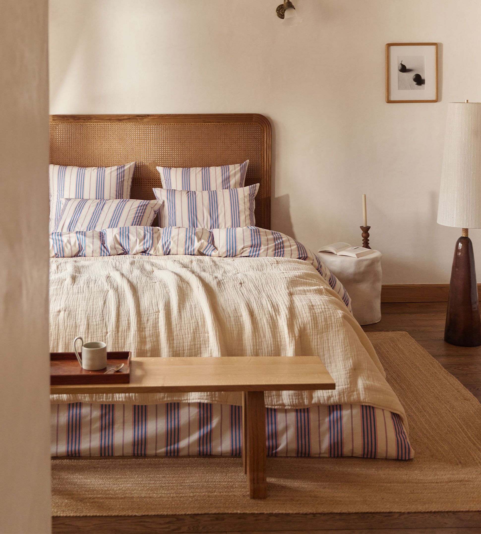 Chambre confortable avec une tête de lit en bois, une literie rayée, un banc en bois, un grand lampadaire, un petit tableau encadré et un tapis tissé.
