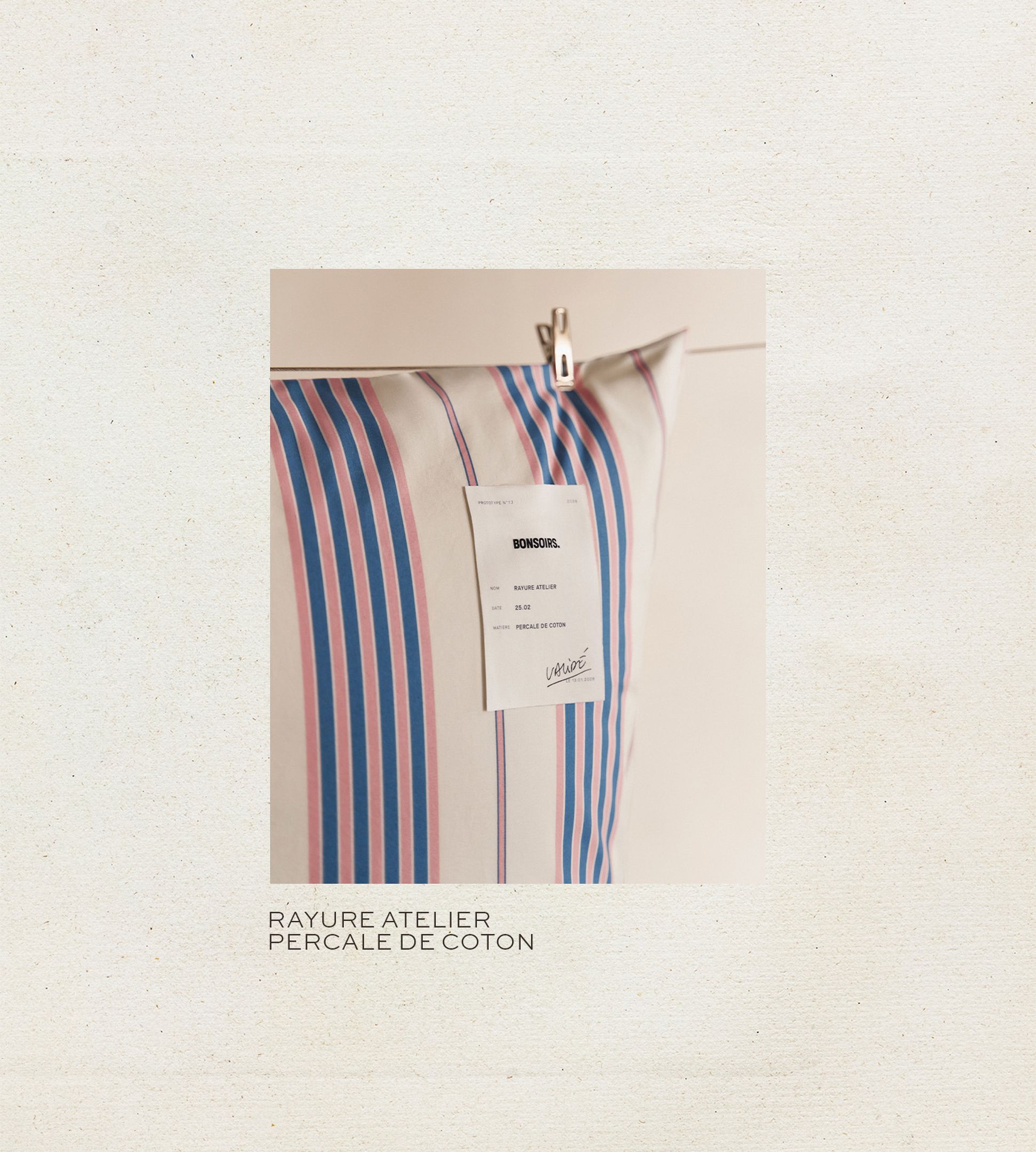 Un oreiller à rayures bleues et roses, orné d'une étiquette blanche portant l'inscription « BONSOIRS », est suspendu à un clip sur un fond clair.