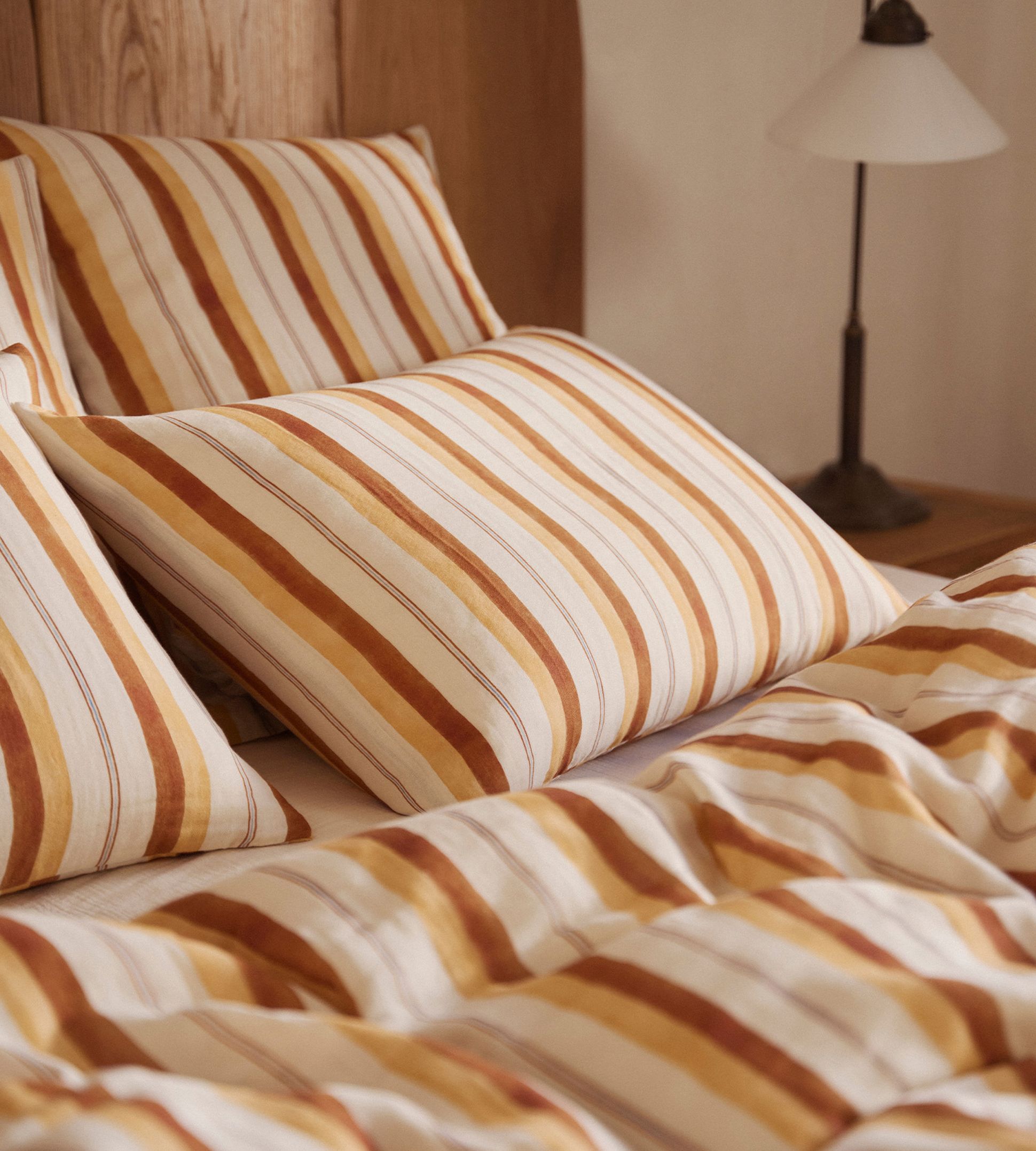 Oreillers rayés et literie aux tons chauds sur un lit en bois, avec une lampe de chevet en arrière-plan.