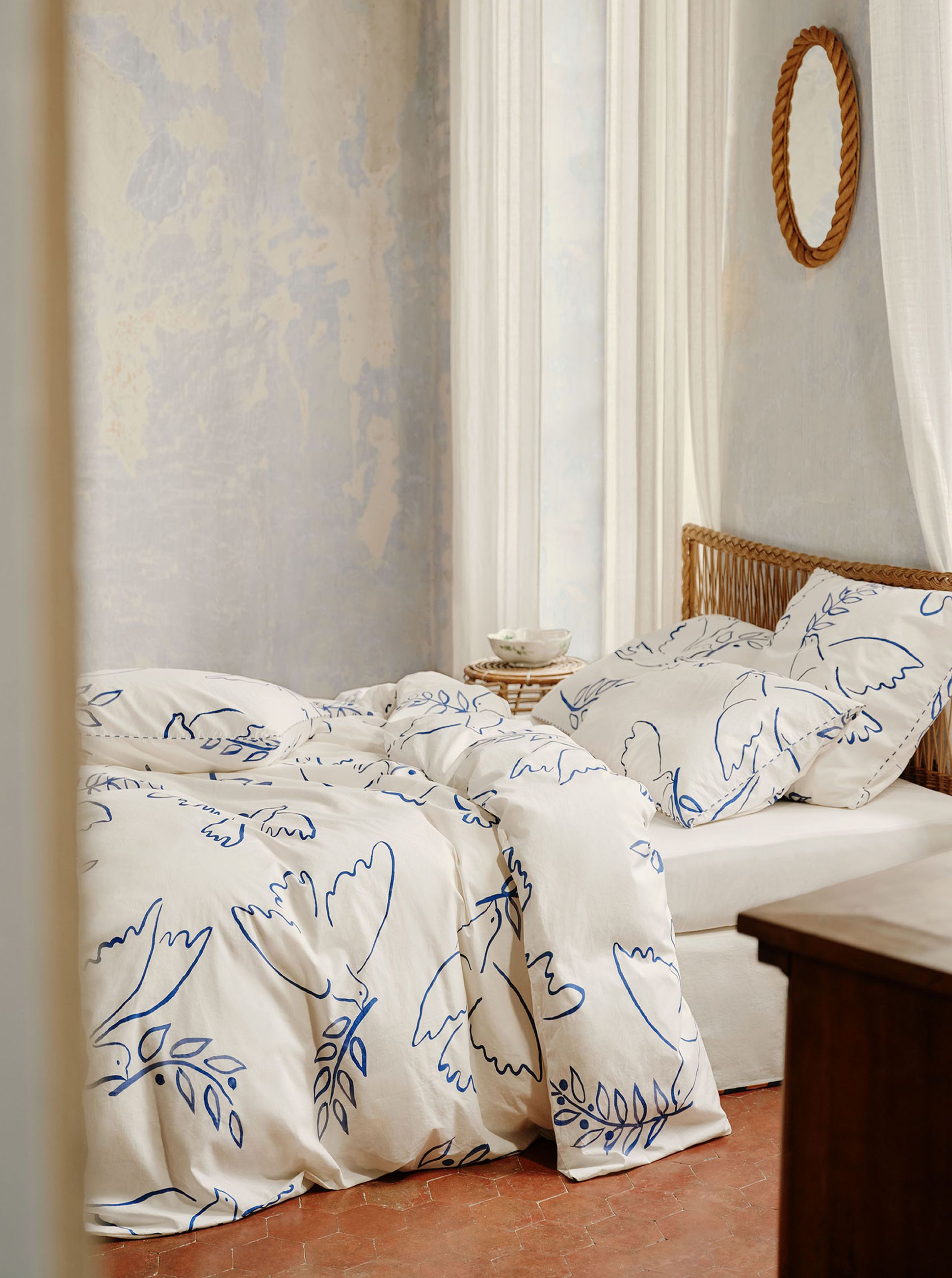 Une chambre douillette avec une parure de lit blanche ornée de motifs botaniques bleus, un lit à cadre en osier, une petite table ronde et un miroir à cadre en corde accroché au mur.