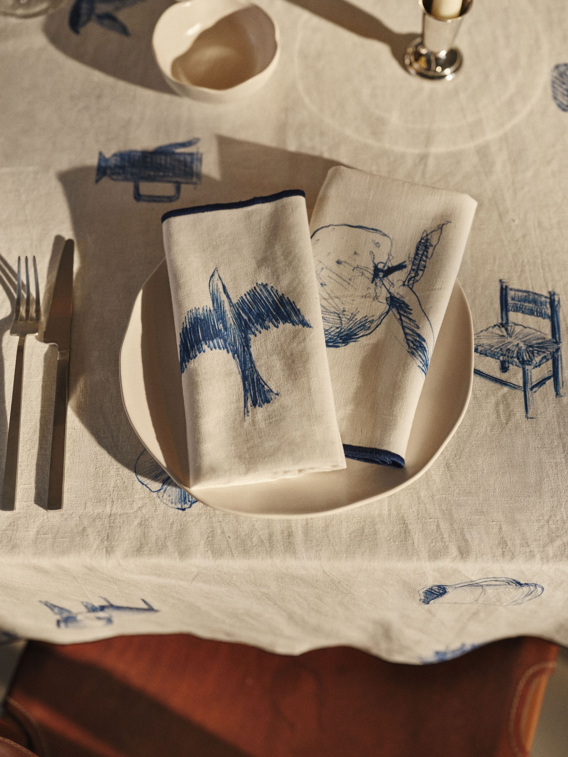 Une table dressée avec deux serviettes illustrées représentant un oiseau et un fruit dans une assiette, posées sur une nappe ornée de croquis bleus similaires.