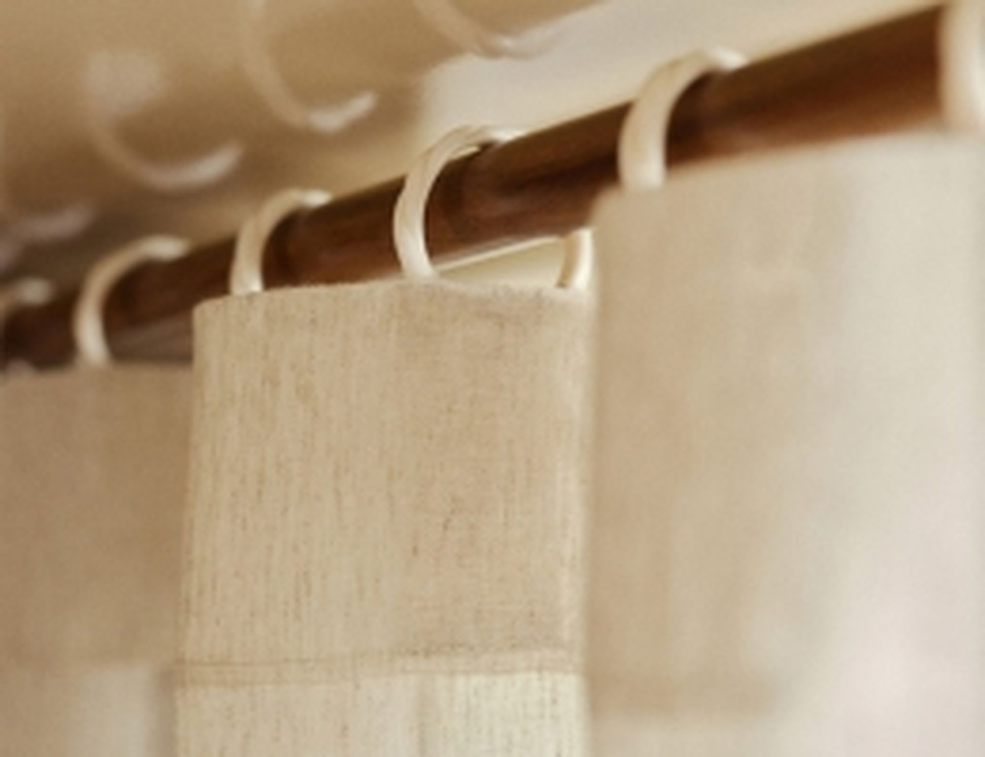 Gros plan sur des rideaux beiges suspendus à une tringle en bois à l'aide d'anneaux en plastique, baignés d'une lumière douce créant une ambiance chaleureuse.