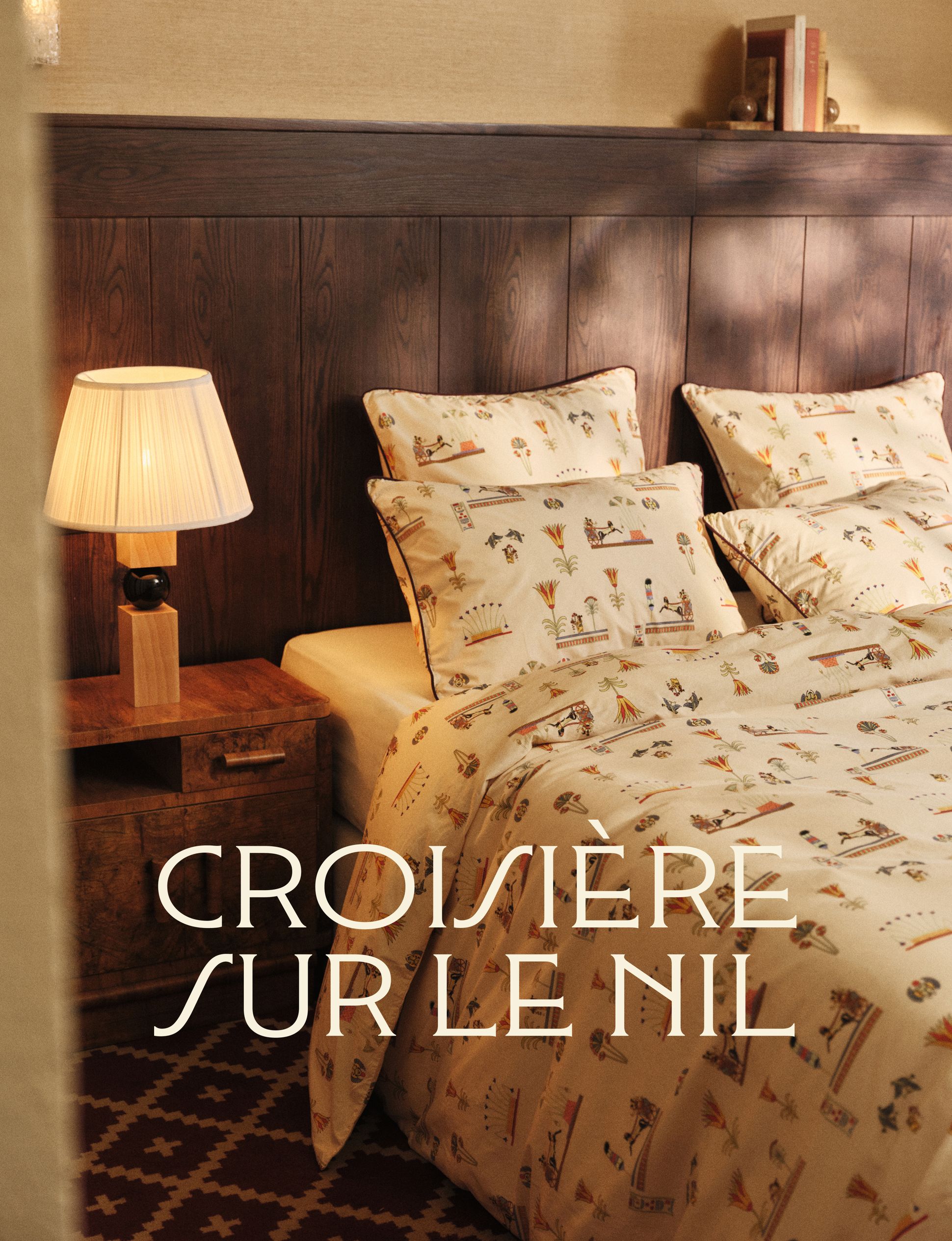 Chambre à coucher chaleureuse avec une parure de lit sur le thème de l'Égypte, une tête de lit en bois, une lampe de chevet et l'inscription « Croisière sur le Nil » superposée.