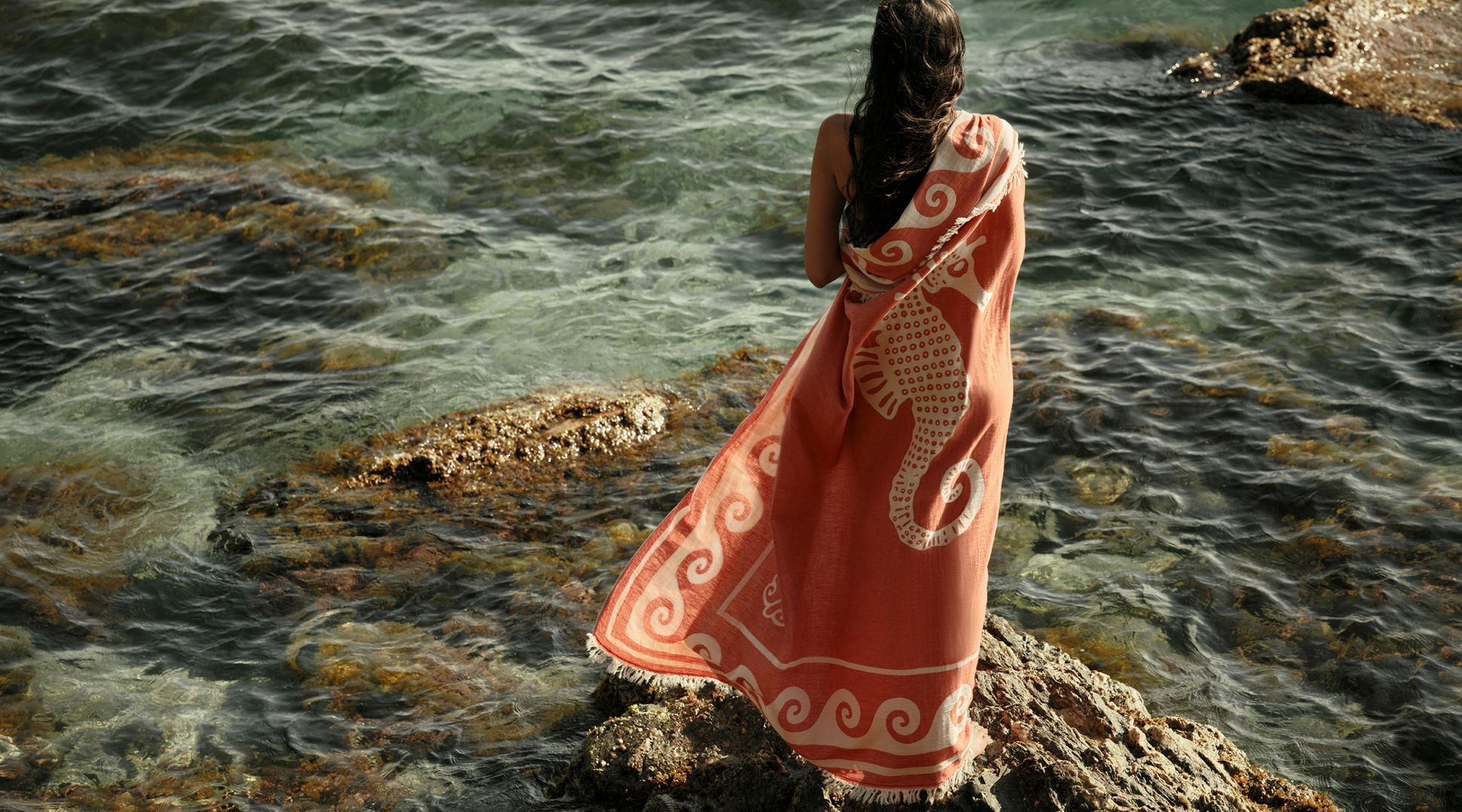 Une personne enveloppée dans une serviette rose ornée d'un motif d'hippocampe, debout sur des rochers au bord de la mer, contemplant l'eau limpide.