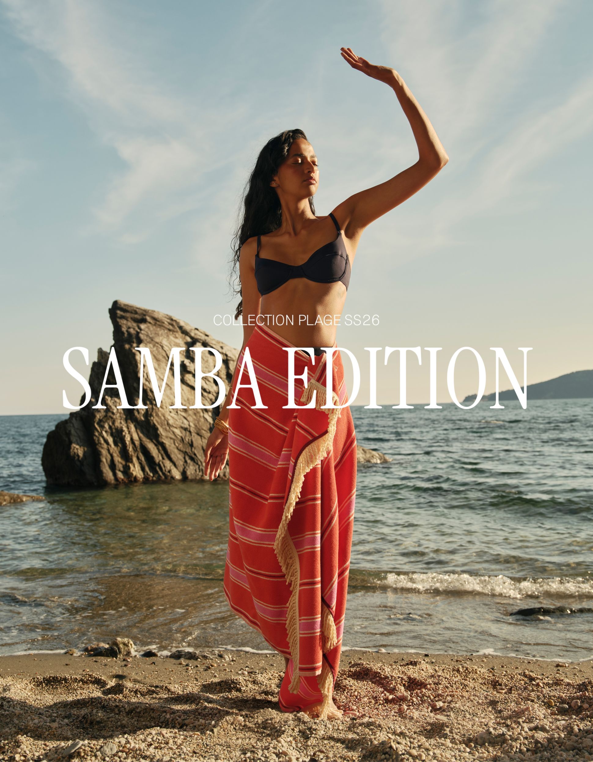 Une femme vêtue de tissus rayés colorés profite de la plage, avec en arrière-plan un rivage rocheux et un ciel dégagé.