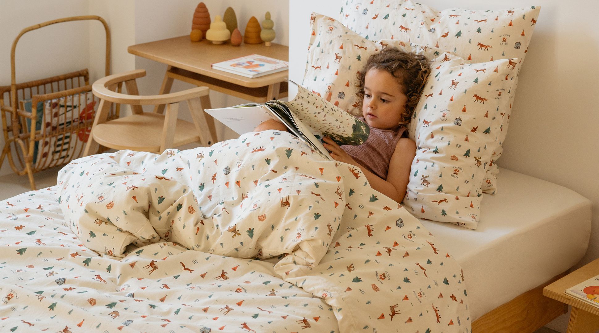 Un enfant lit un livre dans son lit, entouré d'une literie à motifs représentant des arbres et des animaux. Des meubles et des jouets en bois décorent cette chambre confortable.