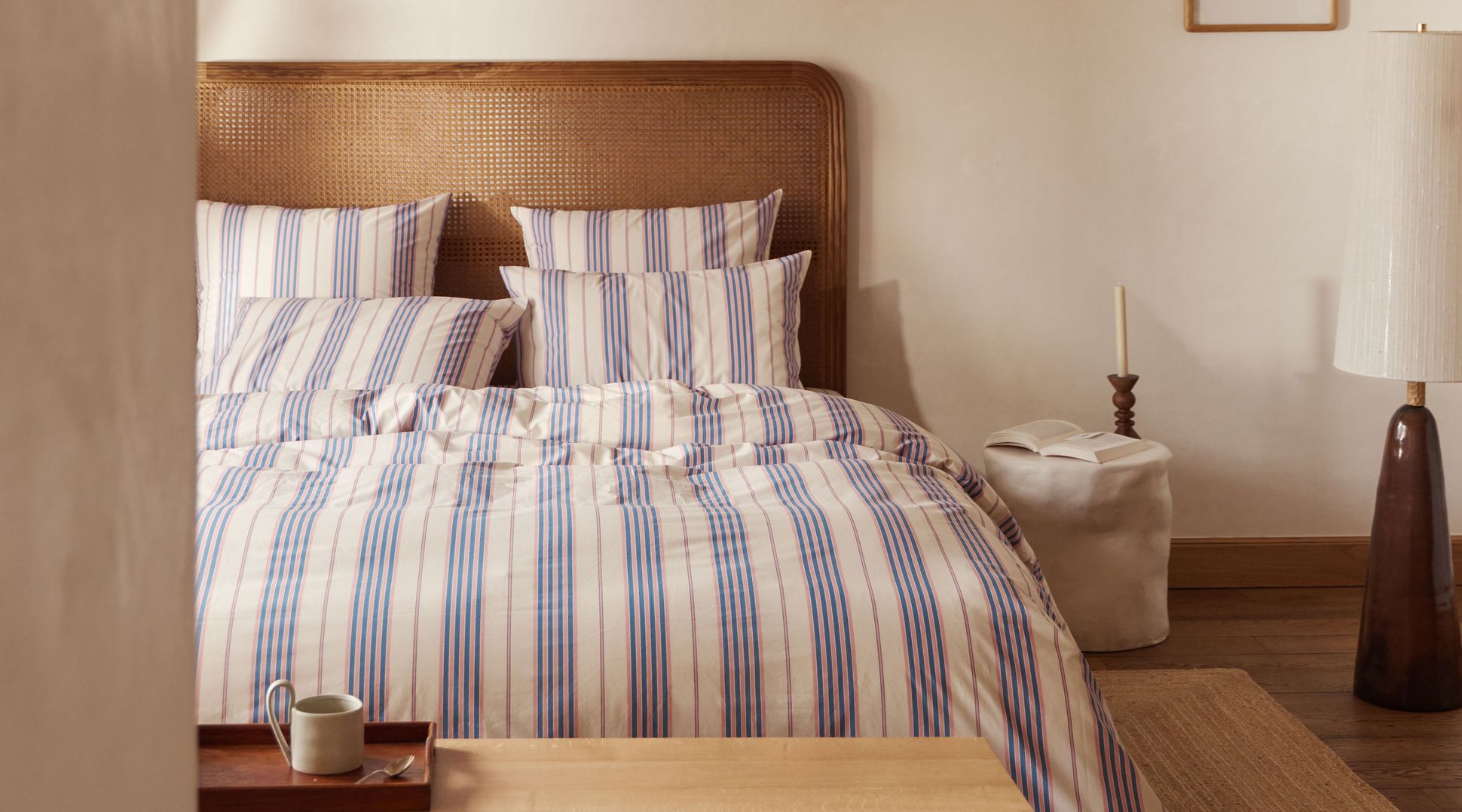 Une chambre douillette avec une parure de lit rayée, une tête de lit en bois et un banc. Une tasse et un plateau sont posés sur le banc, tandis qu'une bougie et une photo trônent sur la table d'appoint.