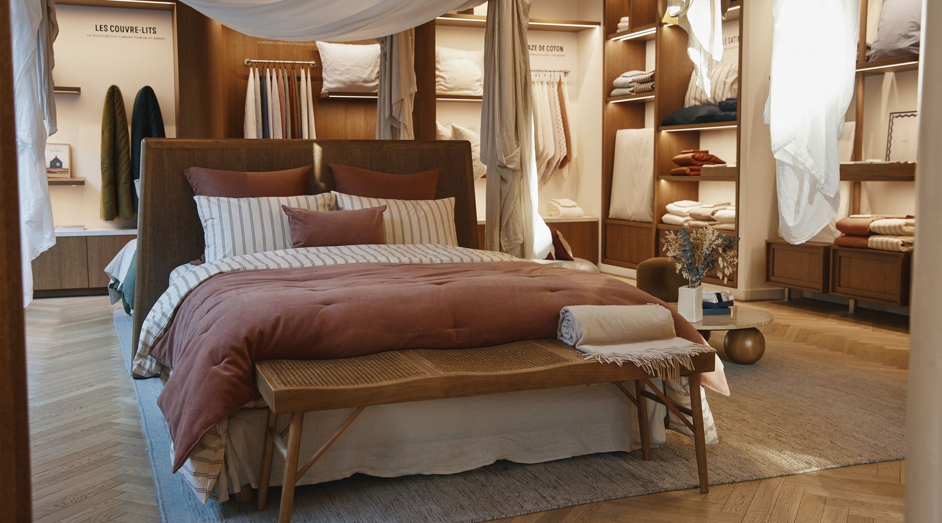 Une chambre à coucher chaleureuse, avec un lit habillé d'une parure à rayures et unise, entouré de tissus drapés et d'étagères en bois sur lesquelles sont disposés des linge de maison.